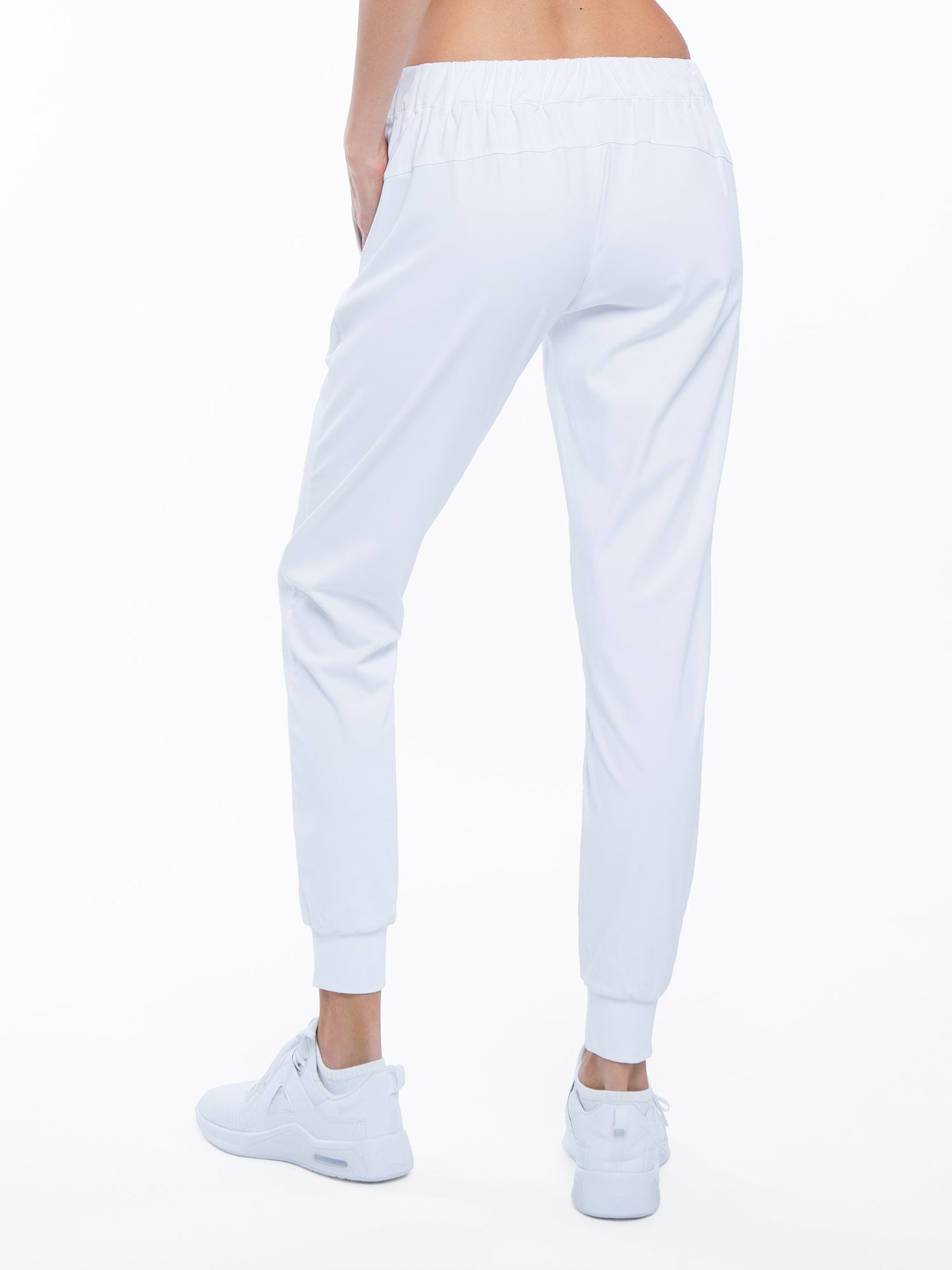 A person is shown from the waist down, facing away, wearing inPhorm NYC’s Alexa Jogger - White with moisture management, elastic waistband and cuffs, paired with white sneakers. The plain white background highlights the clothing.