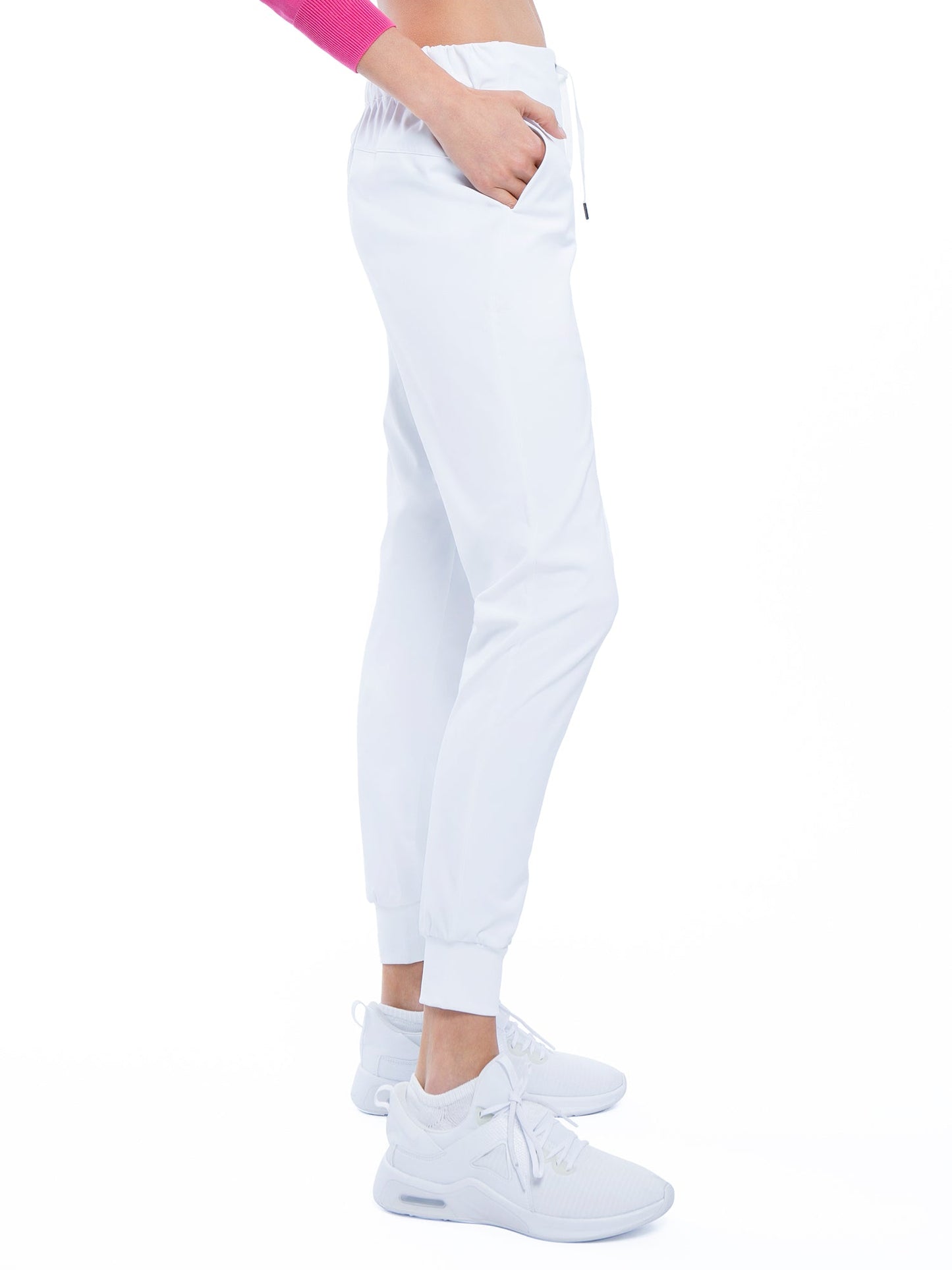 A person stands in profile wearing inPhorm NYC’s Alexa Jogger - White with a pink cropped long-sleeve top and white sneakers, left hand in the pocket, all set against a pure white background for a clean, minimalist look.