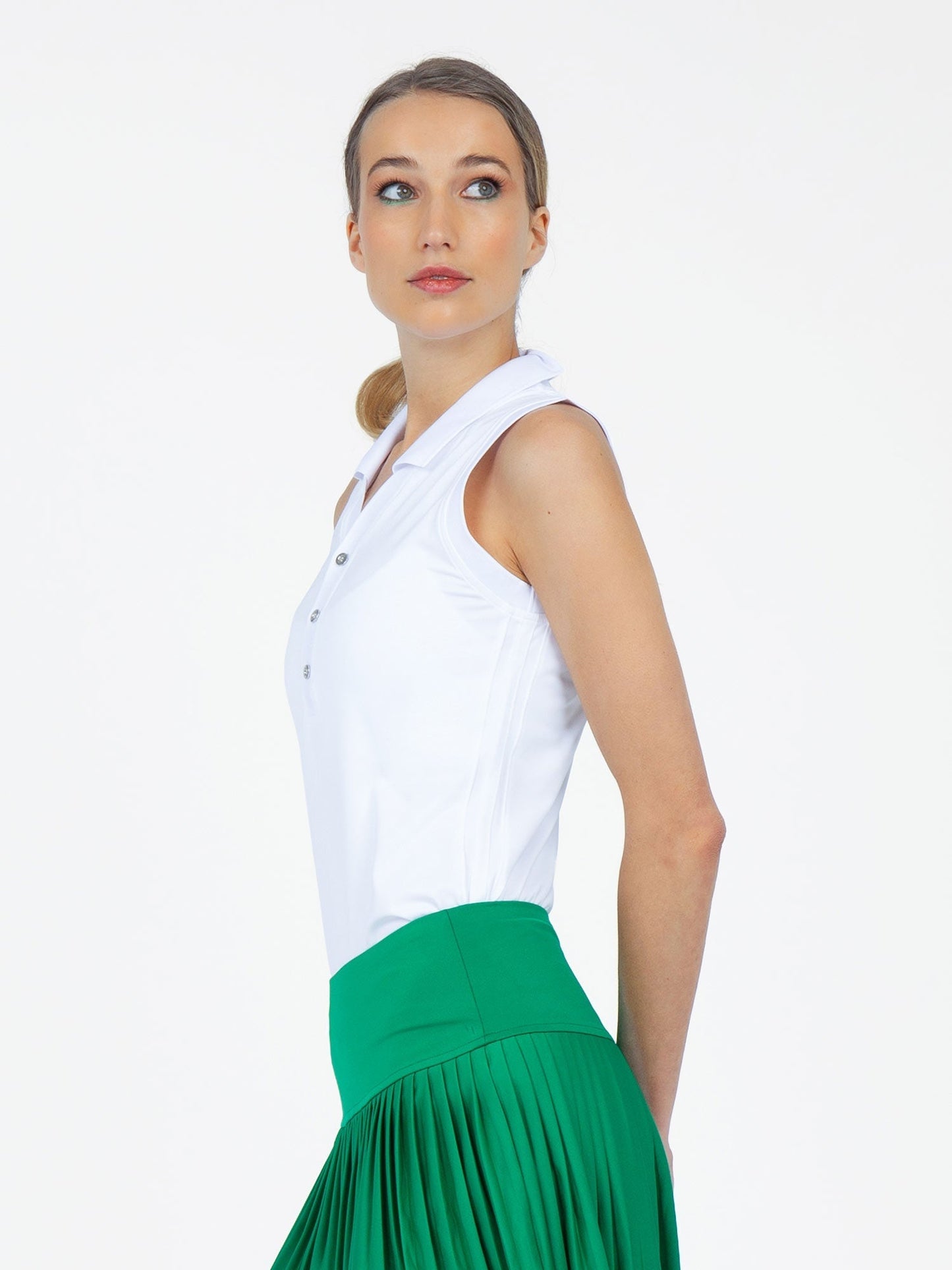 A light-skinned, blue-eyed woman poses sideways, looking over her shoulder. She wears the inPhorm NYC Alexa Performance Sleeveless Polo in White and a high-waisted, pleated green skirt. Her light brown hair is tied back; the background is plain white.