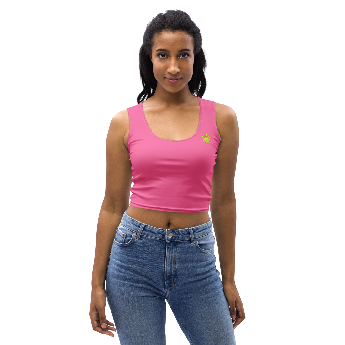 A woman with dark hair faces forward, wearing the Crown Pickleball Women's Corona Crop-top in pink with a small yellow crown graphic and high-waisted blue jeans, standing against a plain white background.