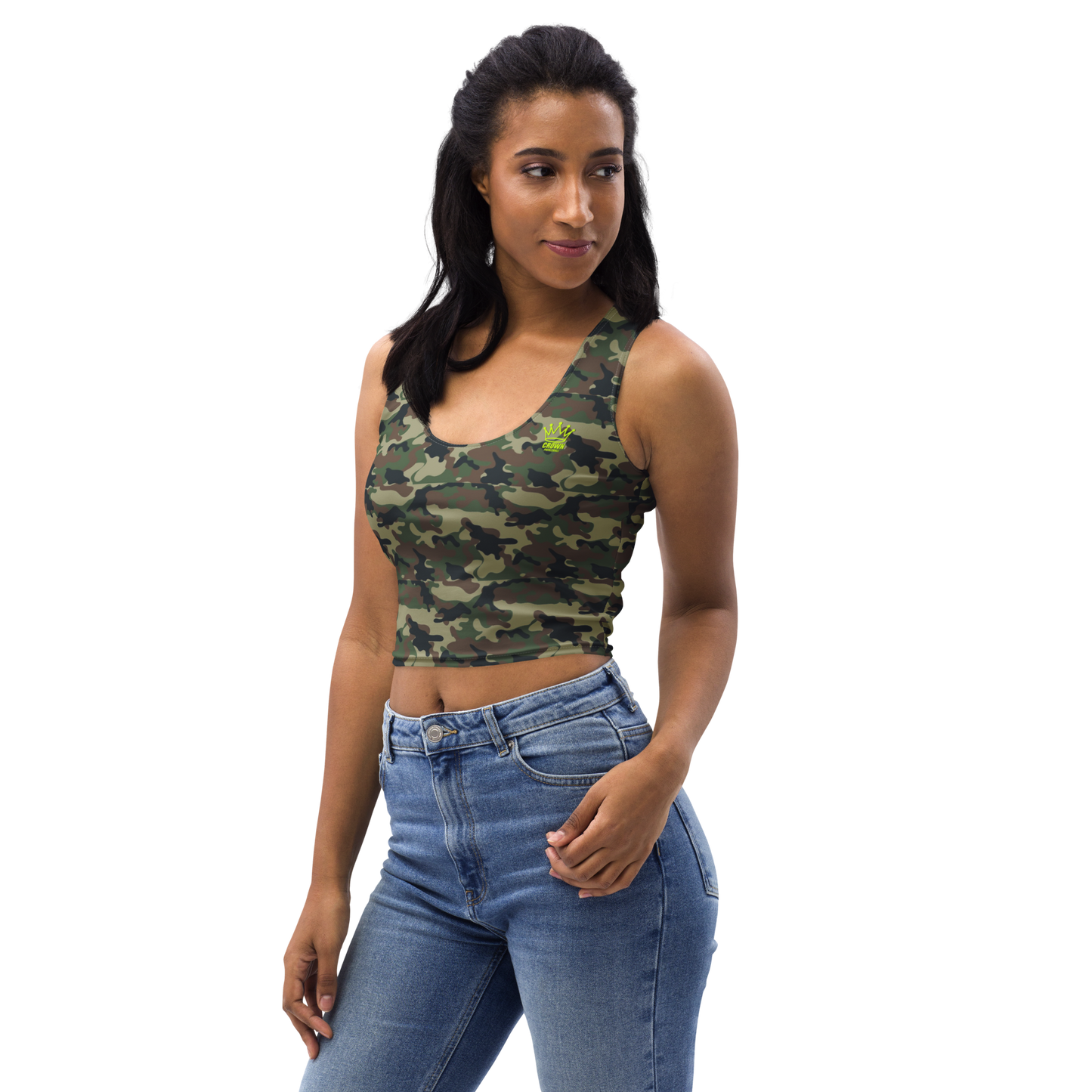 A woman stands against a white background wearing the Crown Pickleball Women's Corona Crop-top (Camo) with blue jeans. Her long dark hair is styled back, and she smiles slightly with one hand in her pocket, looking off to her left.