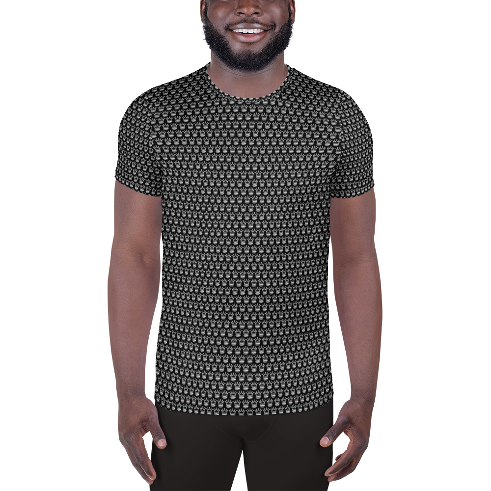 A smiling man with a beard models the Crown Pickleball Men's Corona Pattern Shirt in black, featuring a subtle gray geometric design, as he stands against a plain white background with his arms relaxed at his sides.