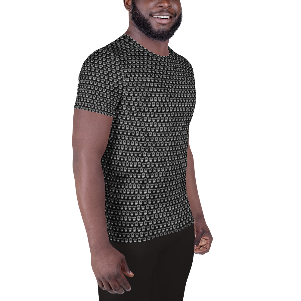 A smiling bearded man stands angled to the side against a white background, wearing the Crown Pickleball Men's Corona Pattern Shirt (Black) with black pants. His left arm hangs relaxed, while his right hand rests in a loose fist.