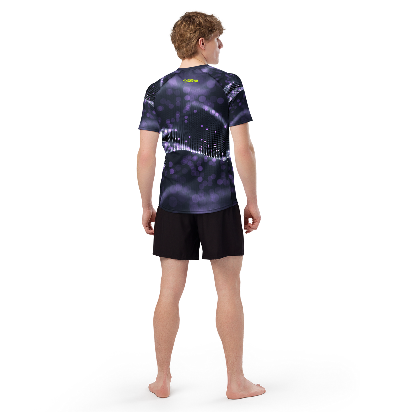 A barefoot person with short light brown hair faces away, wearing the Crown Pickleball Men's Tourney Shirt (Purple) featuring a black and purple graphic wave pattern, made from recycled rash guard material, and black shorts. Plain white background.
