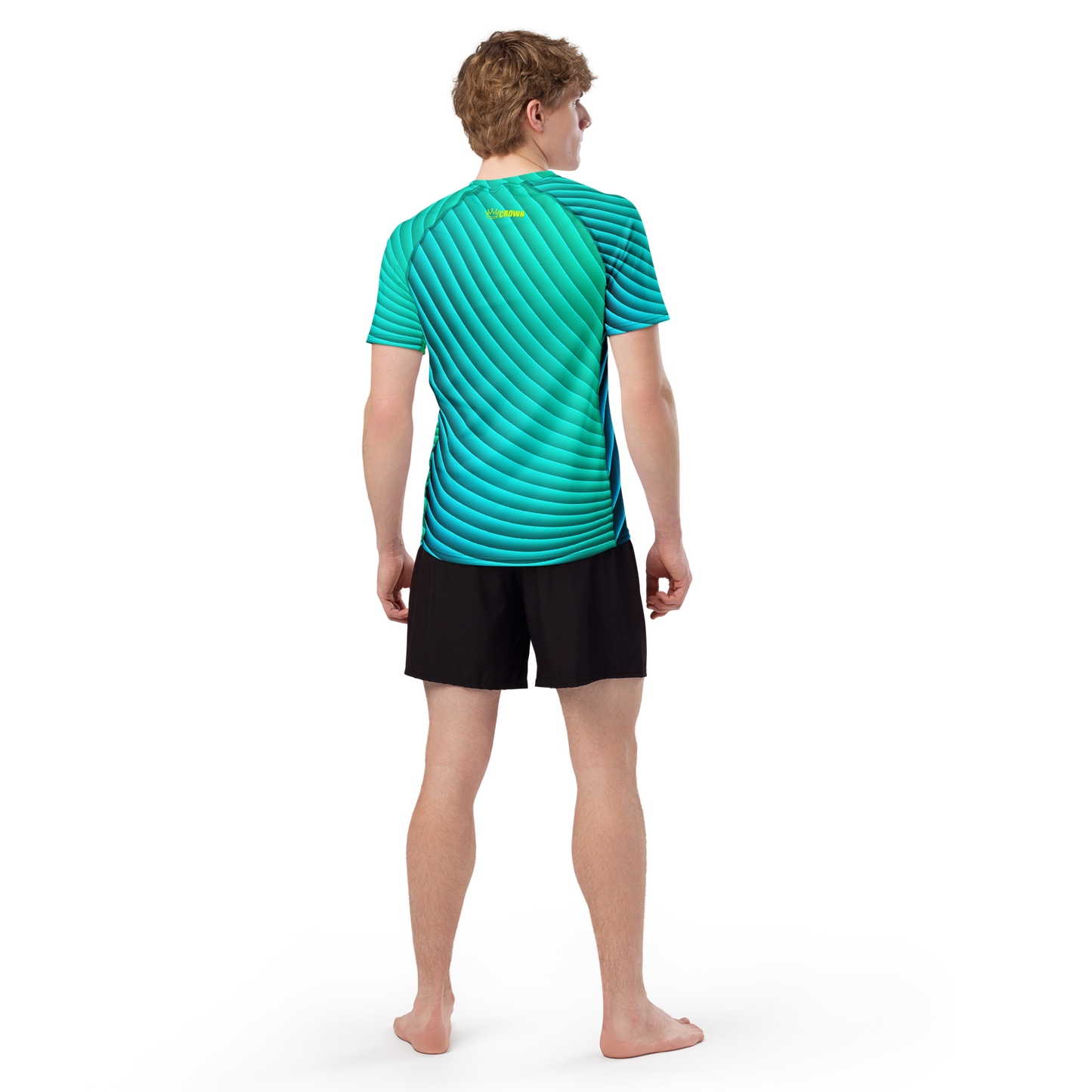 A person with light brown short hair stands barefoot, facing away, wearing the Crown Pickleball Men's Tourney Shirt in turquoise with geometric patterns and black shorts. Arms rest by their sides; background is plain white.