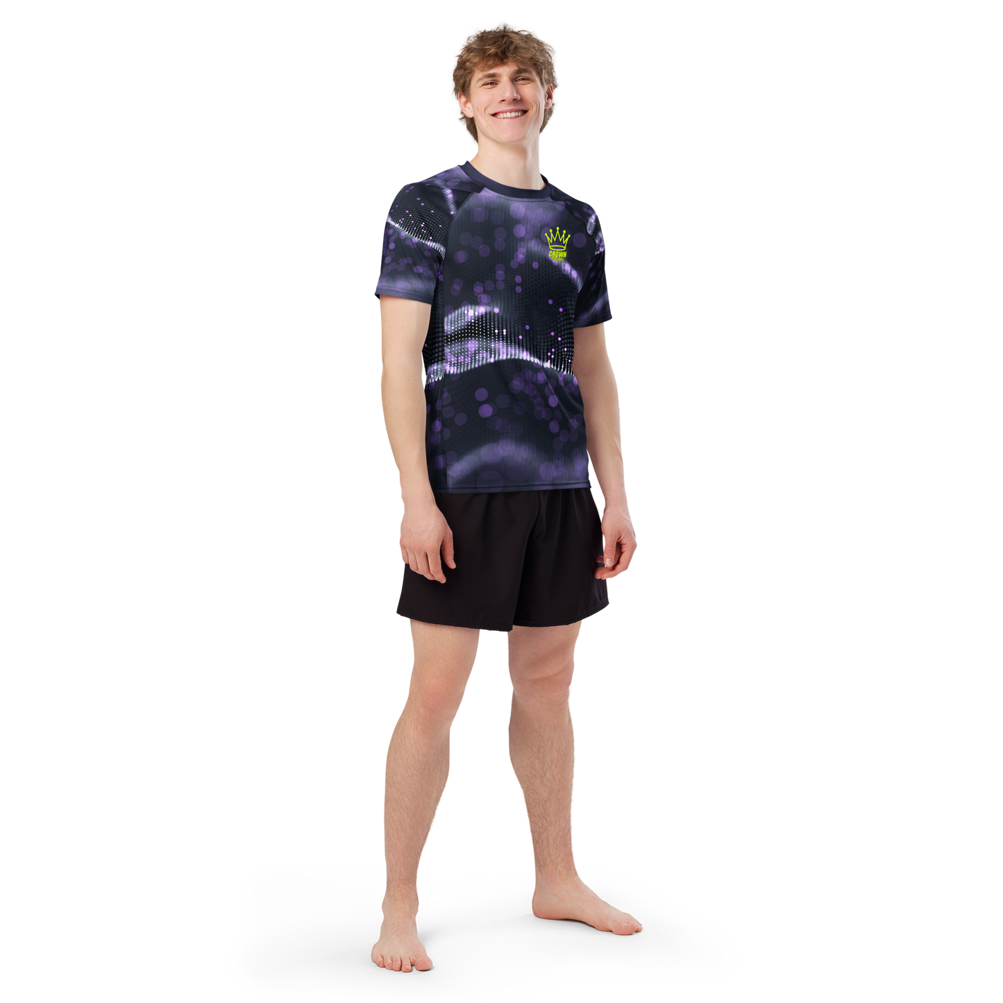 A young man stands barefoot, smiling, wearing the Crown Pickleball Men's Tourney Shirt (Purple) with a green crown logo and black shorts. The moisture-wicking tee by Crown Pickleball is featured against a plain white background, arms relaxed at his sides.