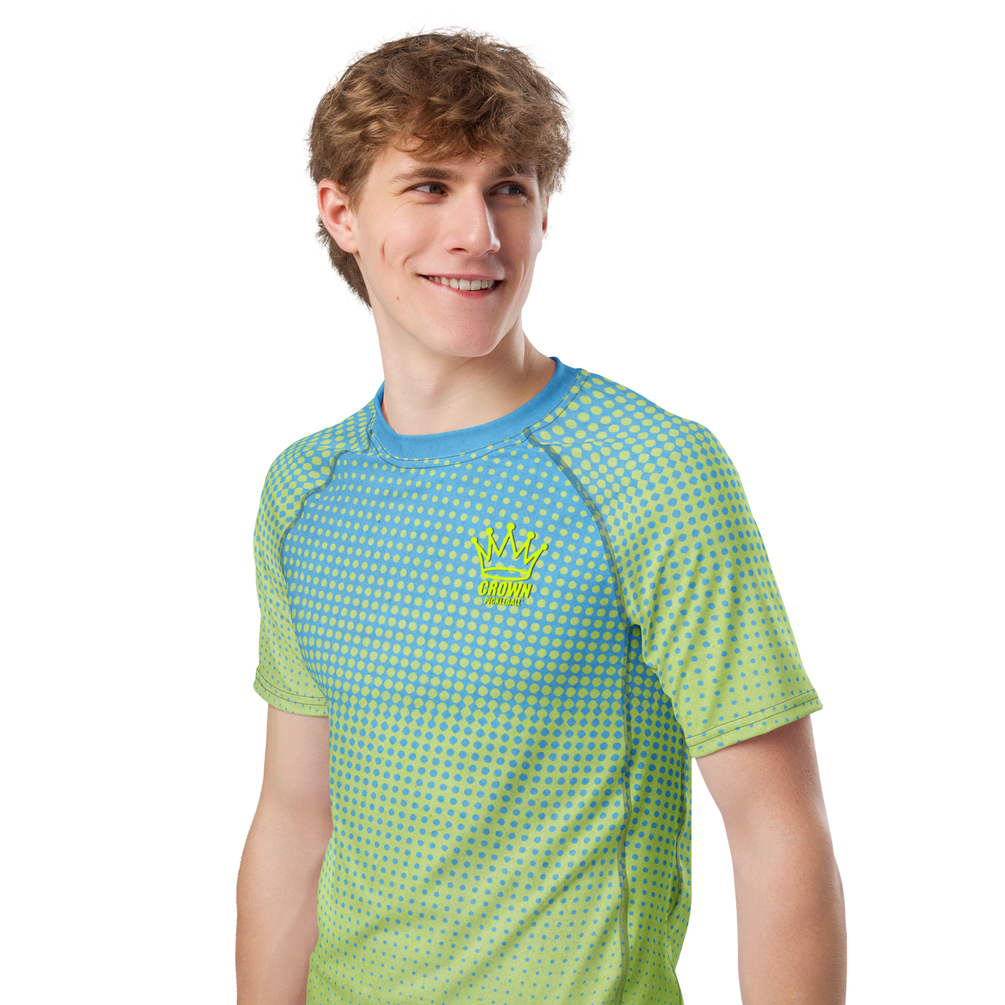 A young man with light brown hair smiles and looks left, wearing the Crown Pickleball Men's Tourney Shirt (Sea)—a blue-green gradient tee with a yellow crown logo, "CROWN" on the chest, dotted pattern, and moisture-wicking fabric.