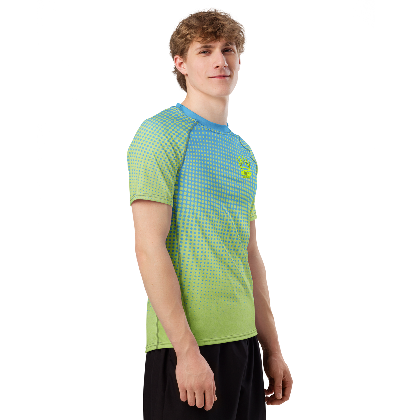 A young man with short, light brown hair stands against a white background, wearing the Crown Pickleball Men's Tourney Shirt (Sea) and black shorts. He faces slightly to the side, smiling softly with one arm relaxed at his side.