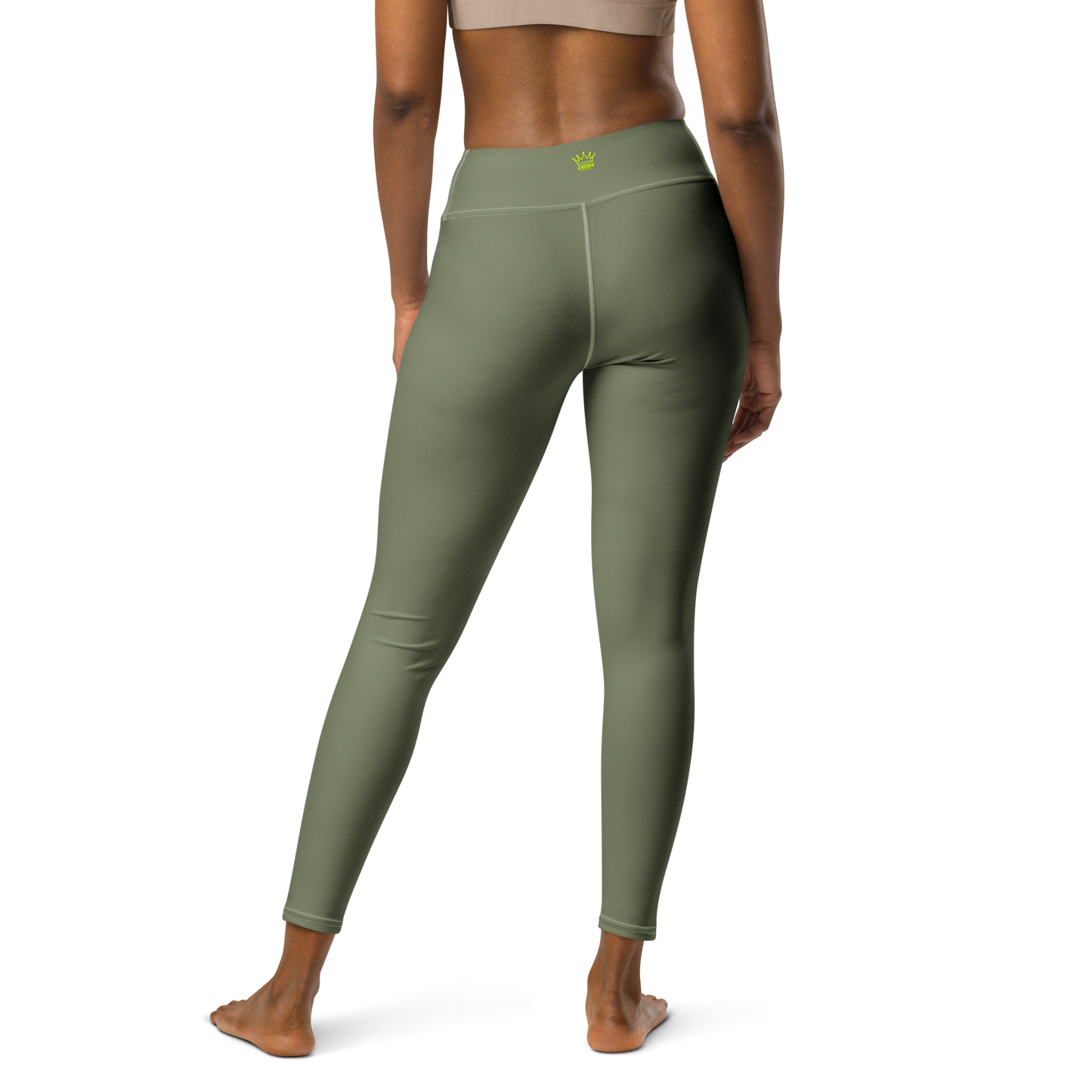 A woman stands barefoot, facing away, wearing Crown Pickleball High-performance Leggings in olive with a yellow crown logo on the high waistband and a beige sports bra. The background is plain white.
