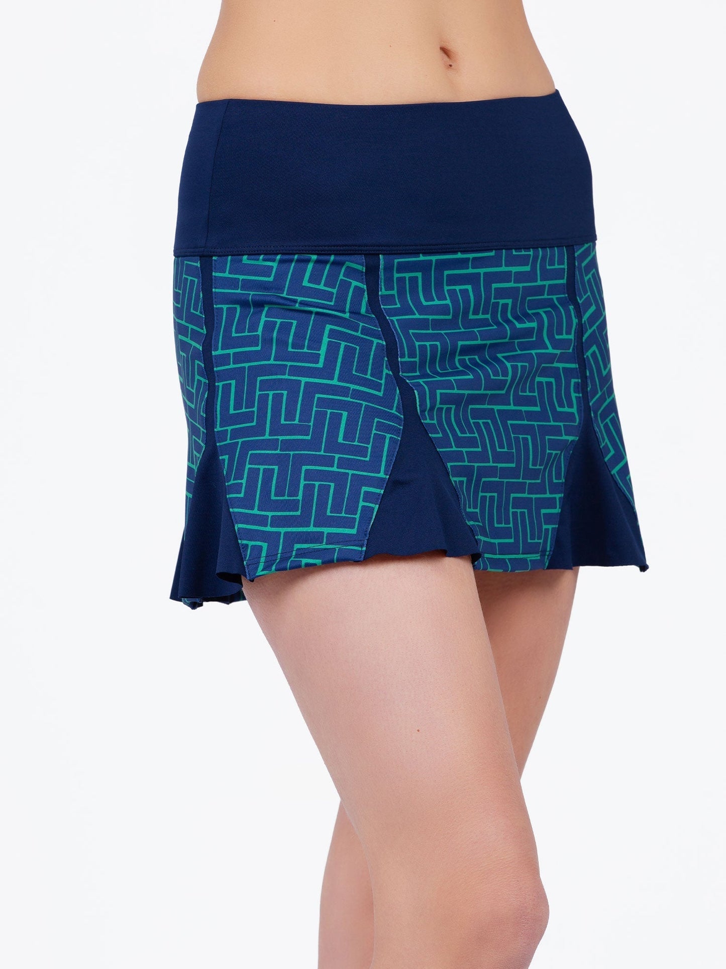 A woman models the Alyssa A-Line Eco-Friendly Skort by inPhorm NYC in Ink Tessel/Ink, featuring a wide waistband, geometric turquoise patterns, two front pleats, and a mid-thigh length. The plain white background highlights the garment.