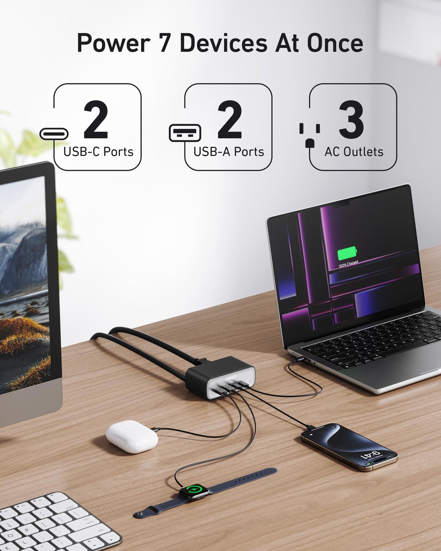 An Anker 7-in-1 USB-C Charging Station (100W) BOA sits on a wooden desk, charging a laptop, phone, earbuds, smartwatch, and more. “Power 7 Devices At Once” is shown above. A monitor appears in the background; three AC outlets are included.