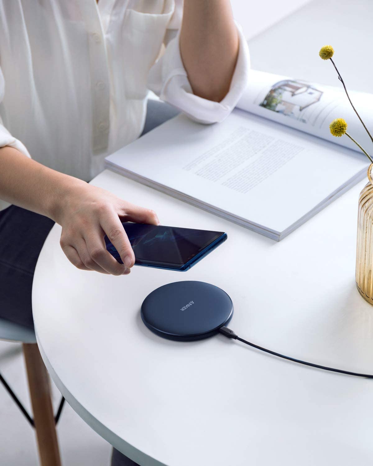 A person in a white shirt holds a smartphone above an Anker 313 Wireless Charger (Pad) by Anker on a round white table, with an open book and yellow flowers in a glass vase creating a calm, modern workspace.