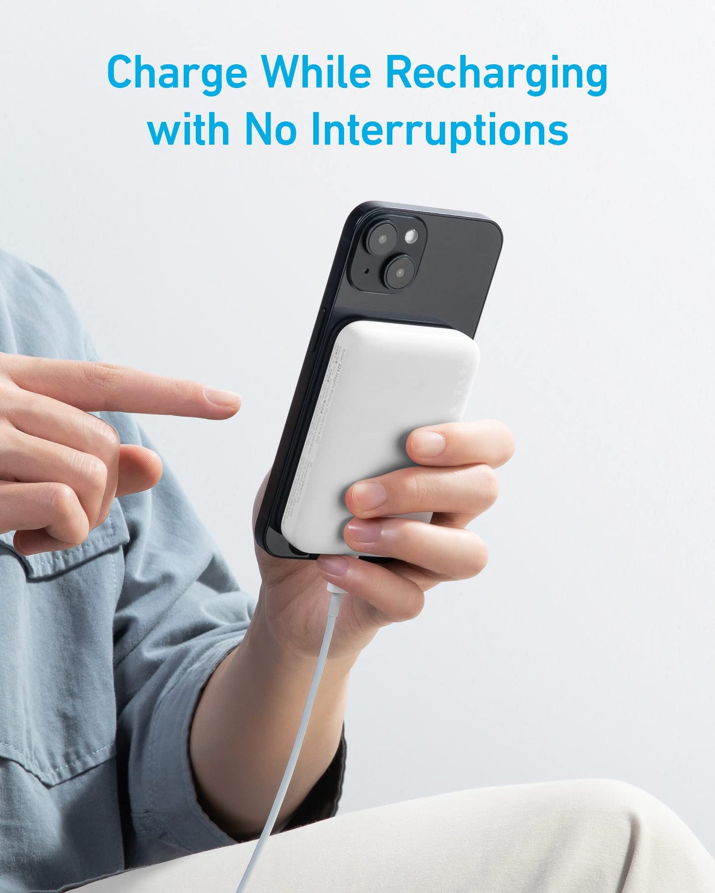 A person holds a black smartphone with the Anker 321 MagGo Battery (PowerCore Magnetic 5K) by Anker attached and charging via cable. Blue text above says: “Charge While Recharging with No Interruptions.”.