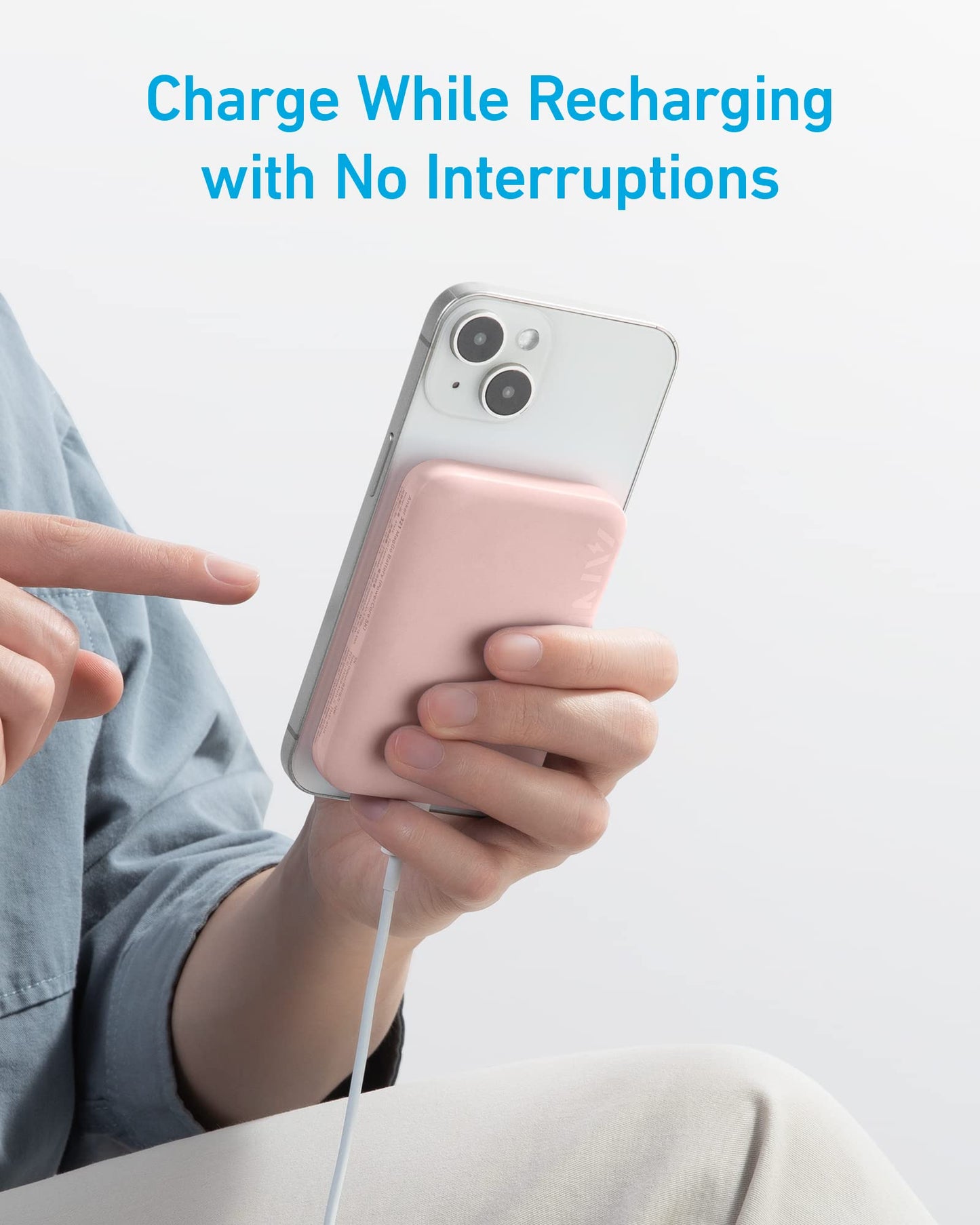A person holds a white smartphone with an Anker 321 MagGo Battery (PowerCore Magnetic 5K) in pink attached to the back via charging cable, pointing at the screen. Blue text above reads: "Charge While Recharging with No Interruptions.