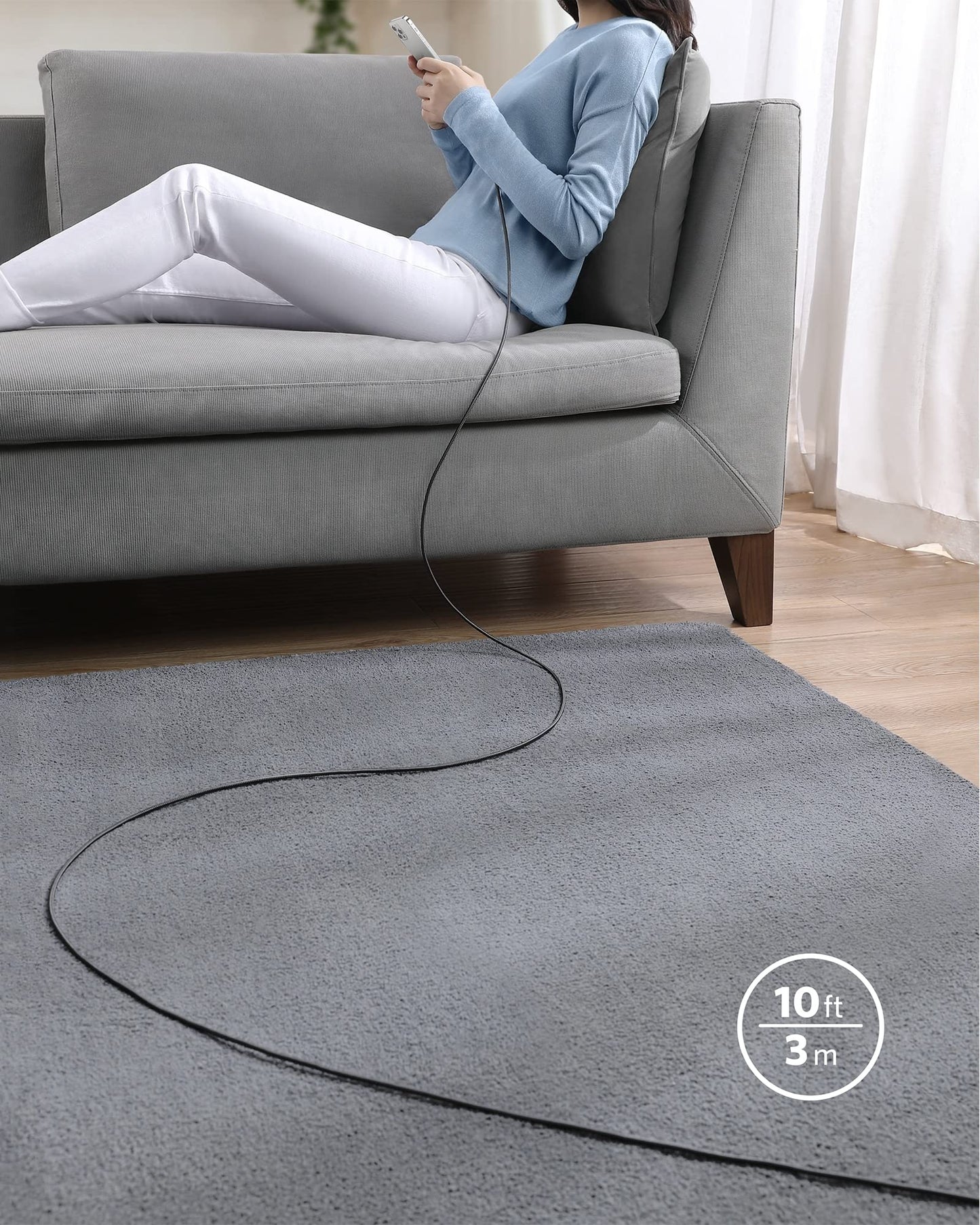 A person sits on a gray sofa using the Anker 331 USB-C to Lightning Cable (MFi Certified, 10 ft) by Anker, charging their iPhone across a gray carpet as sunlight streams through sheer curtains onto the wooden floor.