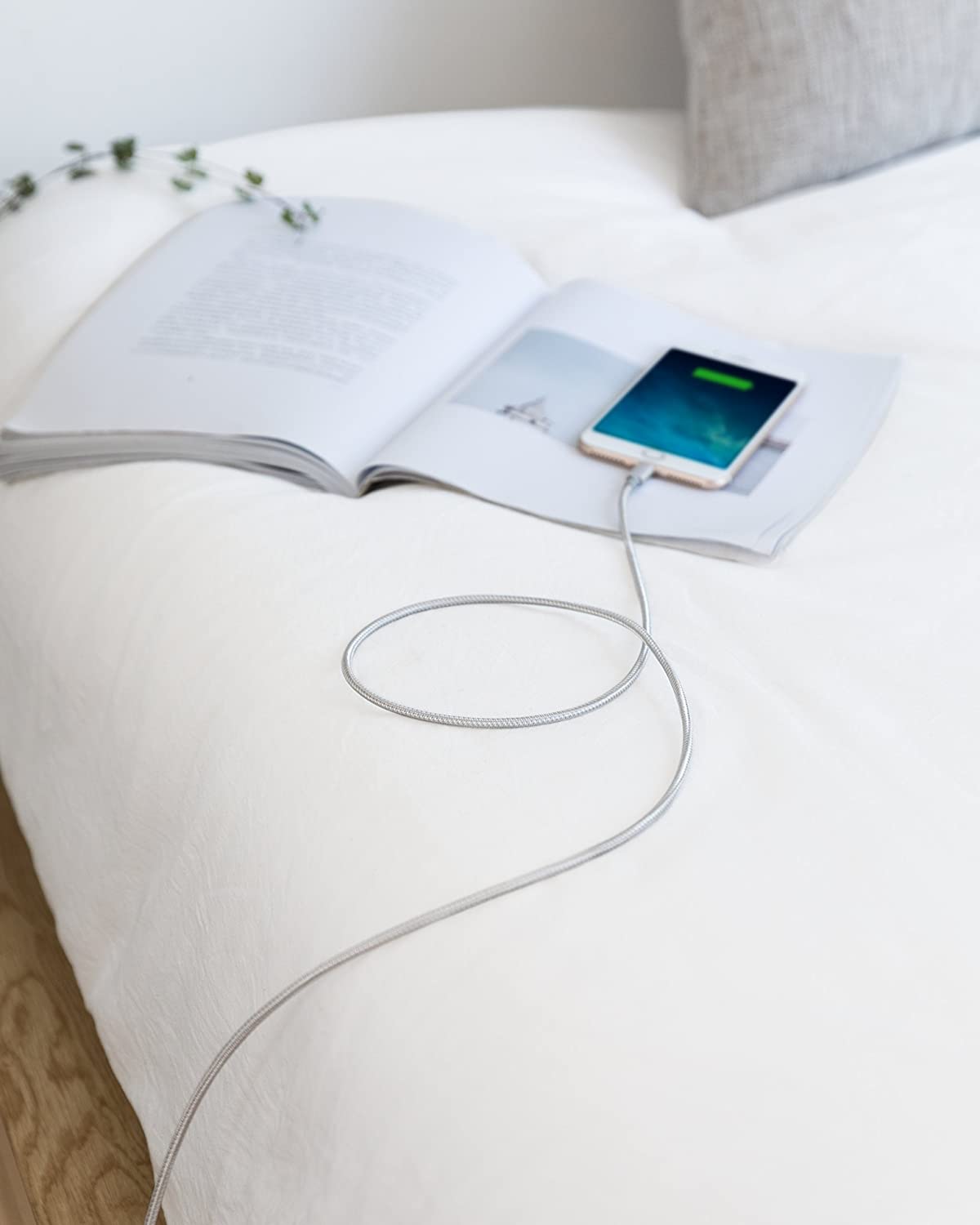 An Anker 331 USB-C to Lightning Cable charges a smartphone resting on an open book atop a white bed. The screen displays a charging icon, while a gray pillow and greenery complete the calm, minimal scene.