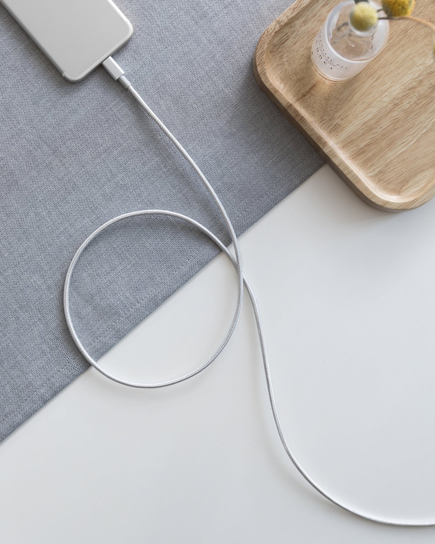 An Anker 331 USB-C to Lightning Cable connects a silver phone on a gray mat atop a white desk. Nearby, a wooden tray holds a glass vase with yellow billy balls and white flowers.