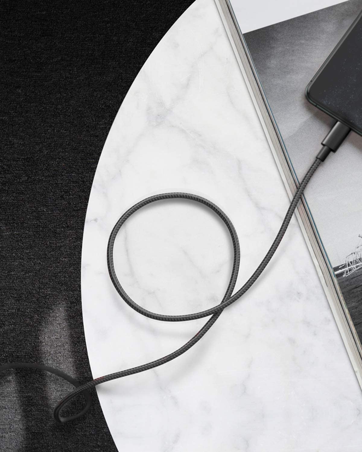 The Anker 331 USB-C to Lightning Cable by Anker forms a loose loop on a white marble surface, connecting to a dark smartphone placed atop an open magazine, with the scene contrasting against a black textured background.