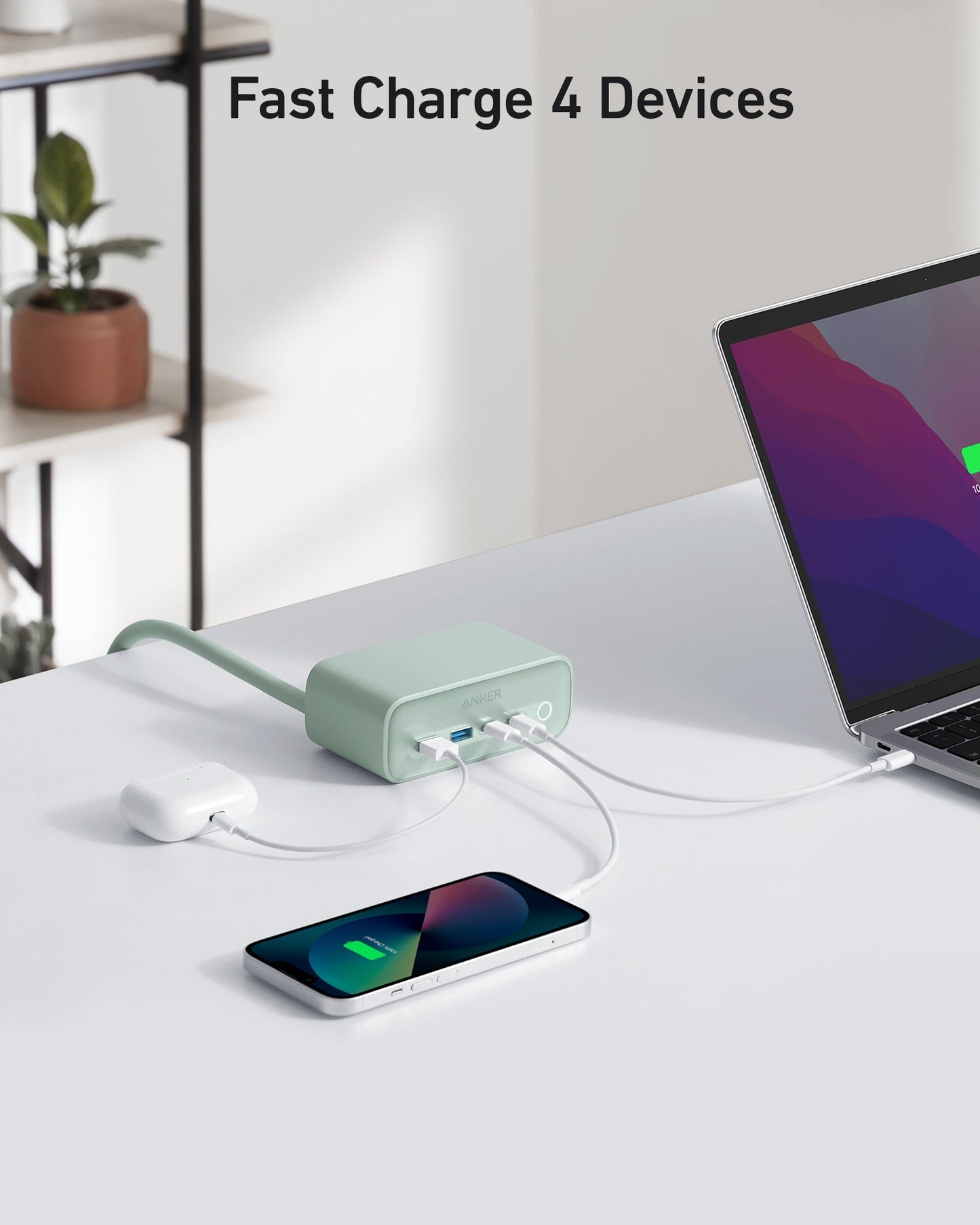 The Anker 525 Charging Station by Anker, shown in green on a white desk, fast-charges a laptop, wireless earbuds, and smartphone. A charging icon is visible on the laptop; the phone lies flat with its indicator lit. Text above reads "Fast Charge 4 Devices.