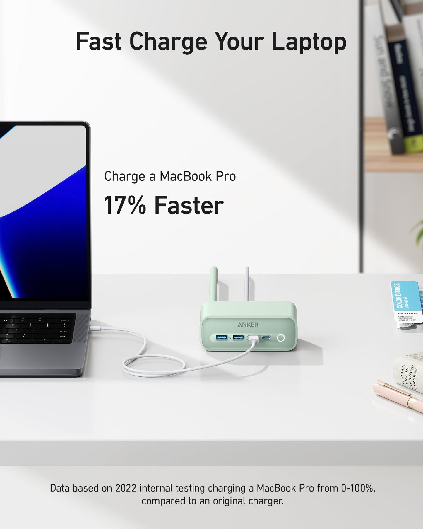 A sleek laptop connects via a white cable to the green Anker 525 Charging Station on a bright desk. Text reads: “Fast Charge Your Laptop. Charge a MacBook Pro 17% Faster.” Books, a plant, and office supplies appear in the background.