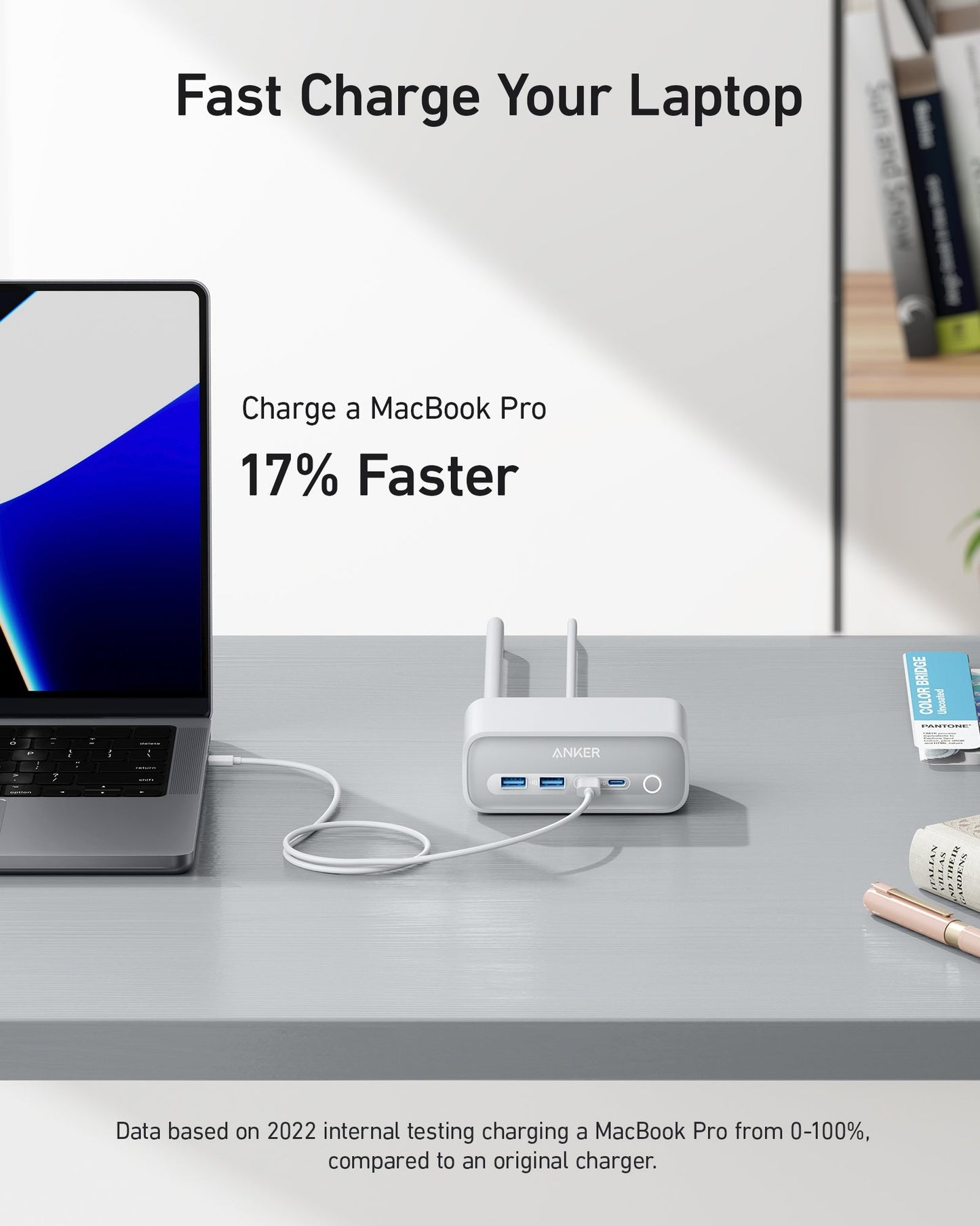 A silver laptop on a gray desk connects via white cable to the gray Anker 525 Charging Station from Anker, featuring two antennas. Text above reads: Fast Charge Your Laptop. Charge a MacBook Pro 17% Faster. A notebook and pen are on the right.