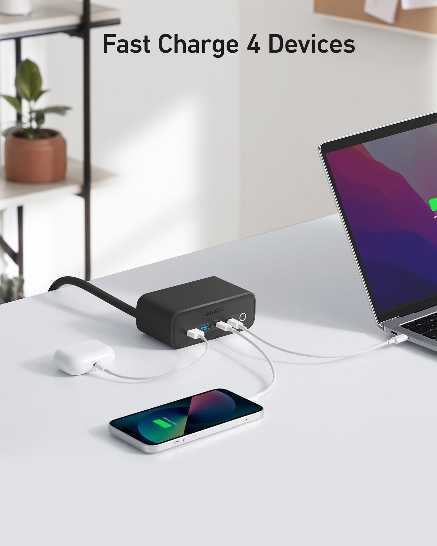 The Anker 525 Charging Station in black sits on a white desk, charging a laptop, wireless earbuds, and a smartphone with white cables. Phone and laptop screens display charging icons. Text above says: "Fast Charge 4 Devices." A plant is nearby.