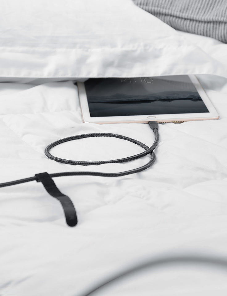 A tablet partly under a white pillow on a white bed is connected to an Anker 551 USB-A to Lightning Cable, with the cable resting on the bedding. A hint of gray blanket appears in the corner and the dim tablet screen displays a faint time.