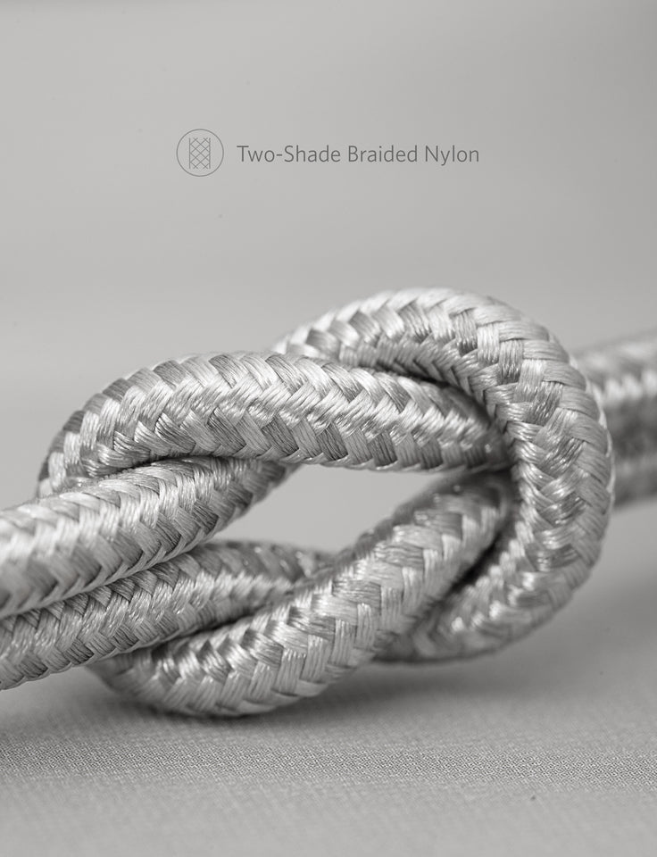 A silvery, two-shade braided Anker 551 USB-A to Lightning Cable—with a visible overhand knot—shows its tightly woven nylon fibers gleaming against a smooth gray background. Text above reads “Two-Shade Braided Nylon.”.