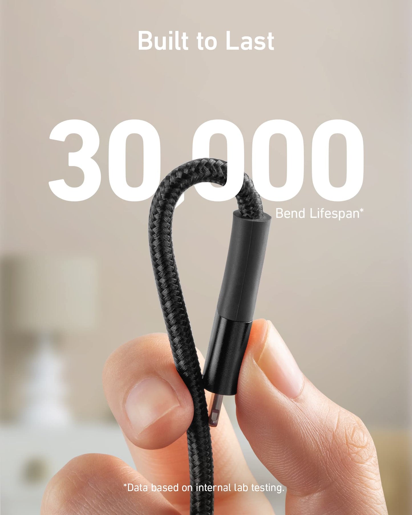 A hand holds a black Anker 551 USB-A to Lightning Cable bent in a U shape, with bold text saying “30,000 Bend Lifespan” and “Built to Last” above. The background is blurred with faint home furnishings visible.