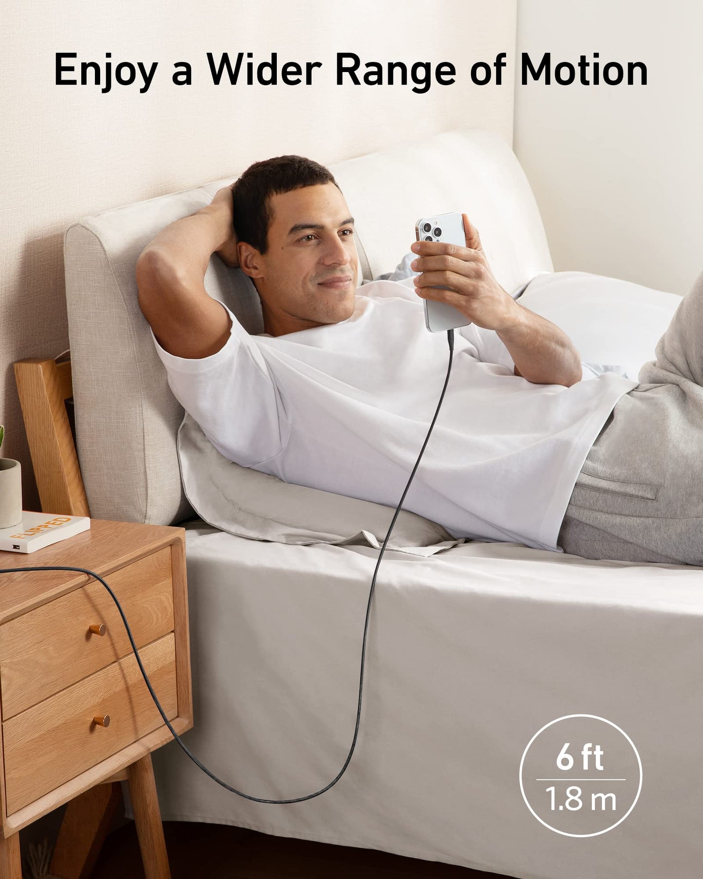 A man reclines on a bed, smiling while using his smartphone connected with the Anker 551 USB-A to Lightning Cable. Text above reads “Enjoy a Wider Range of Motion.” Bottom right: “6 ft | 1.8 m.” Brand: Anker.