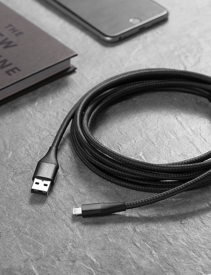 An Anker 551 USB-A to Lightning Cable in black is coiled on a gray stone surface, with a dark-covered book and a smartphone in a black case blurred in the background.