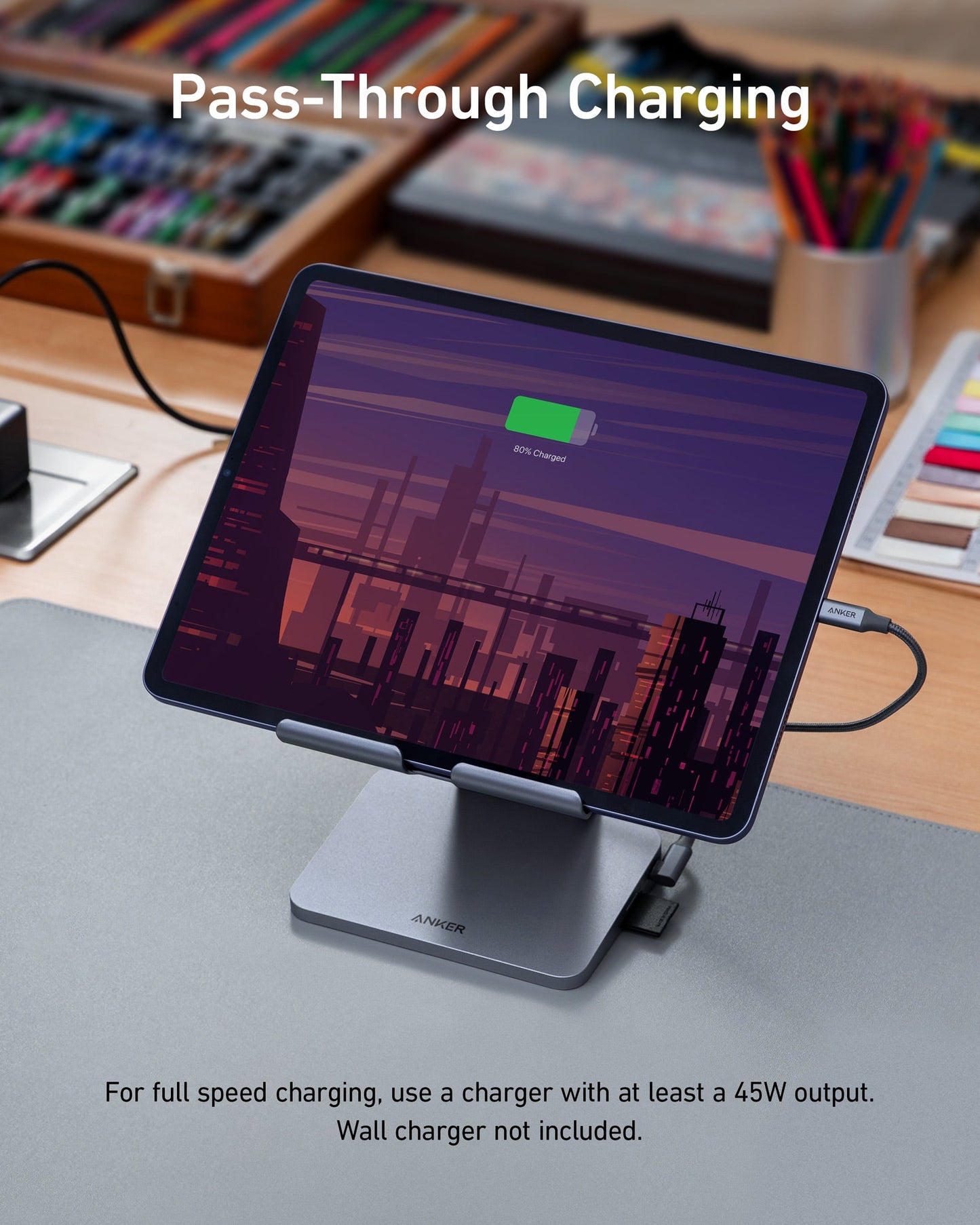 Anker <b>551</b> USB-C Hub (8-in-1, Tablet Stand) holds a tablet at 80% charge, connected to power. Desk has art supplies and books behind. Pass-through charging shown; note: use a 45W charger (not included).