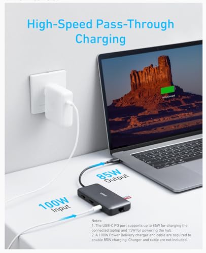 On a white desk, a laptop displays a desert scene while charging at 85W via an Anker <b>553</b> USB-C Hub (8-in-1), which receives 100W from the charger and outputs 85W Power Delivery to the laptop.