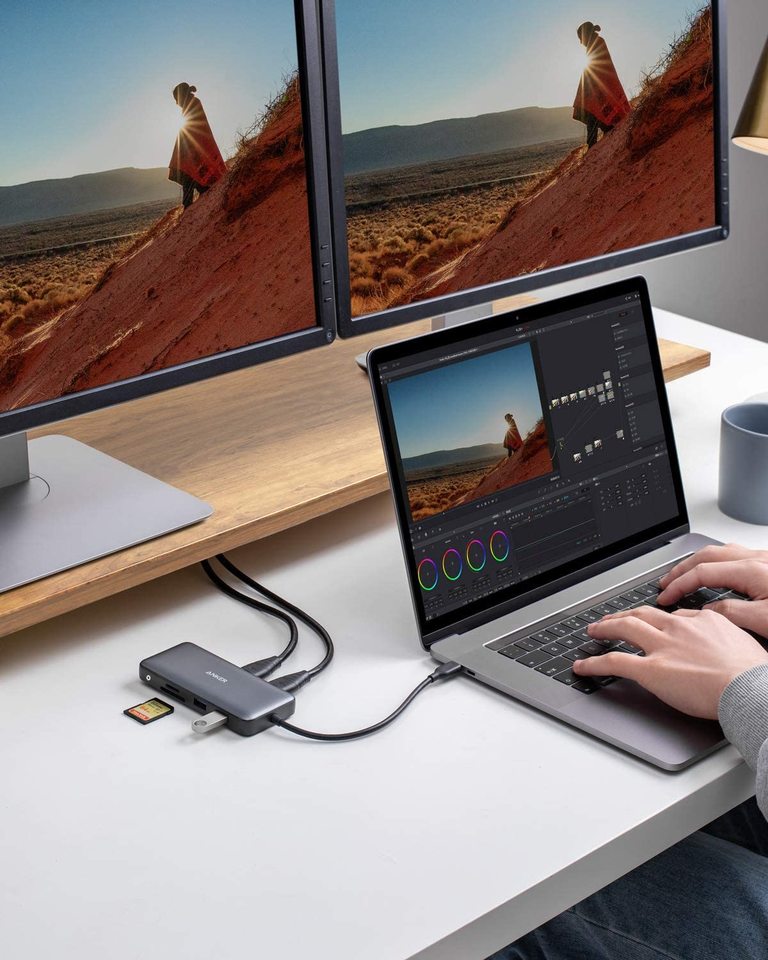 A person uses a laptop connected to the Anker 553 USB-C Hub (8-in-1), displaying scenic desert images on dual HDMI monitors. Multiple cables and SD cards are plugged in, with a mug and mouse on the white desk for a complete tech setup.