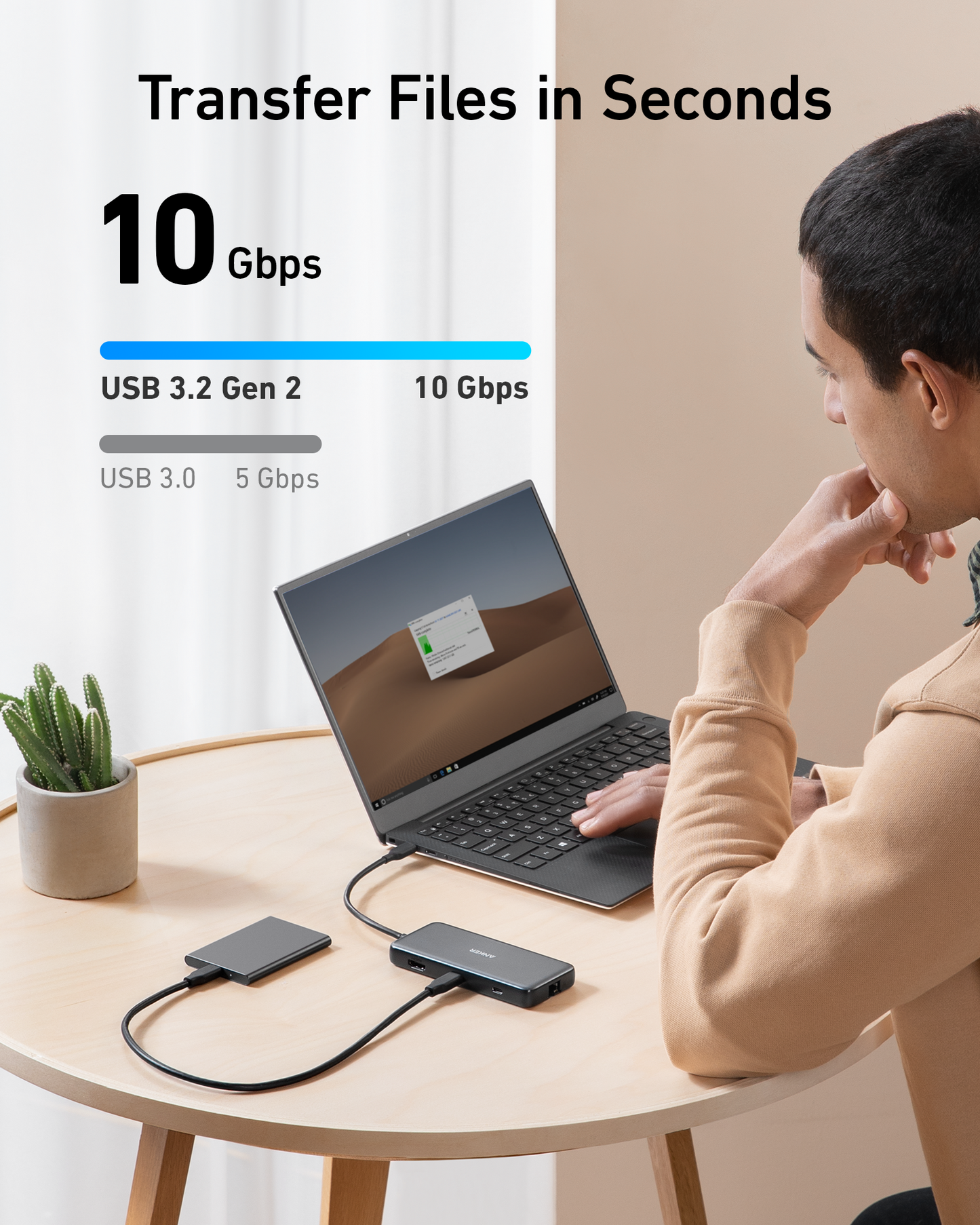 A person uses a laptop with an Anker <b>555</b> USB-C Hub (8-in-1) and external hard drive at a round wooden table, featuring Power Delivery. A cactus pot decorates the scene as graphics compare USB 3.0 (5Gbps) vs USB 3.2 Gen 2 (10Gbps) speeds.
