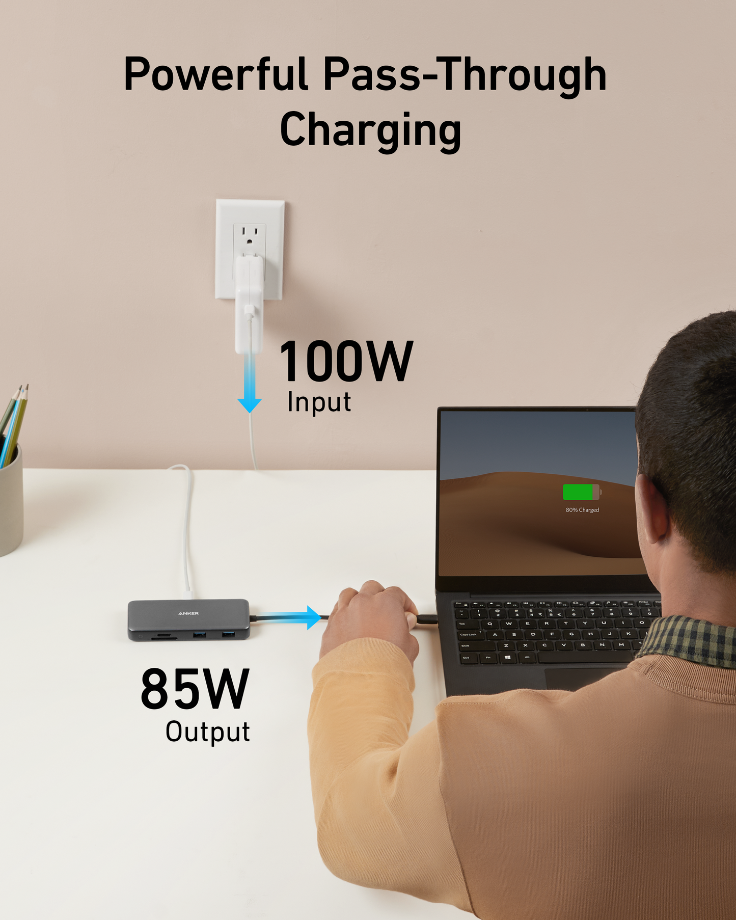 A person uses a laptop at a desk, connected to an Anker 555 USB-C Hub (8-in-1). Plugged into a wall outlet labeled “100W Input,” the hub delivers “85W Output” and powerful Power Delivery charging to the laptop.