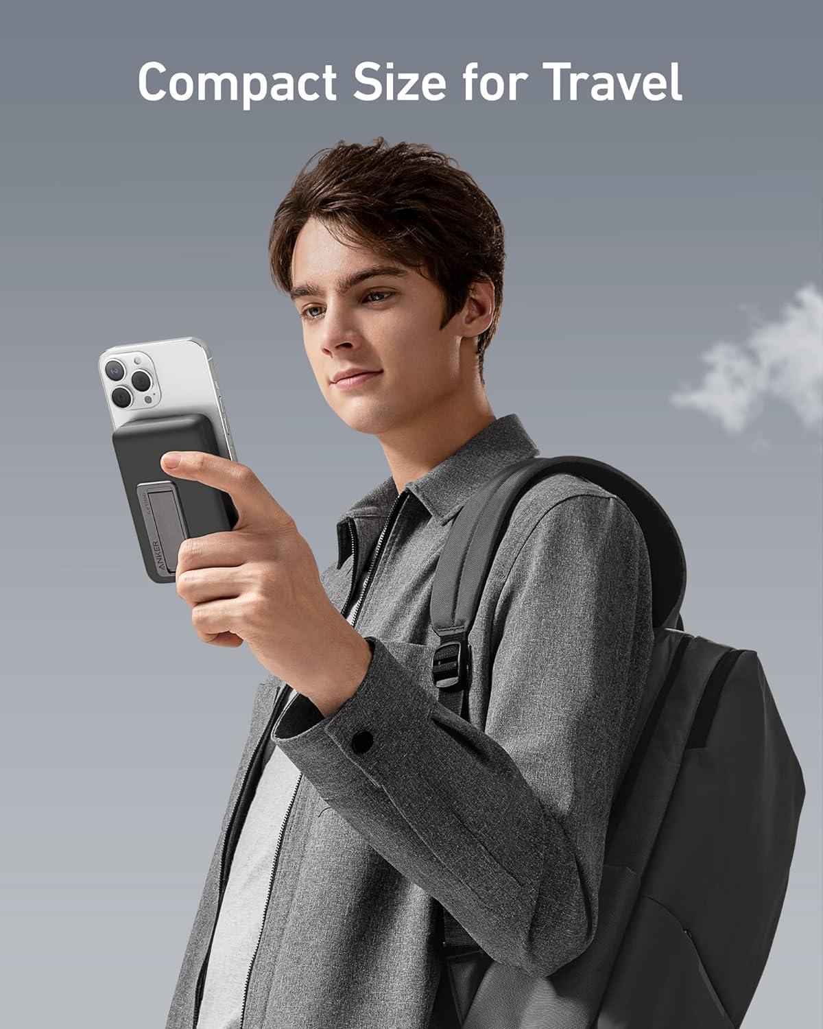 A young man with brown hair, in a grey shirt and backpack, holds his phone with the Anker 633 Magnetic Battery by Anker attached. He looks at his phone against a pale sky backdrop. Text above reads: Compact Size for Travel.