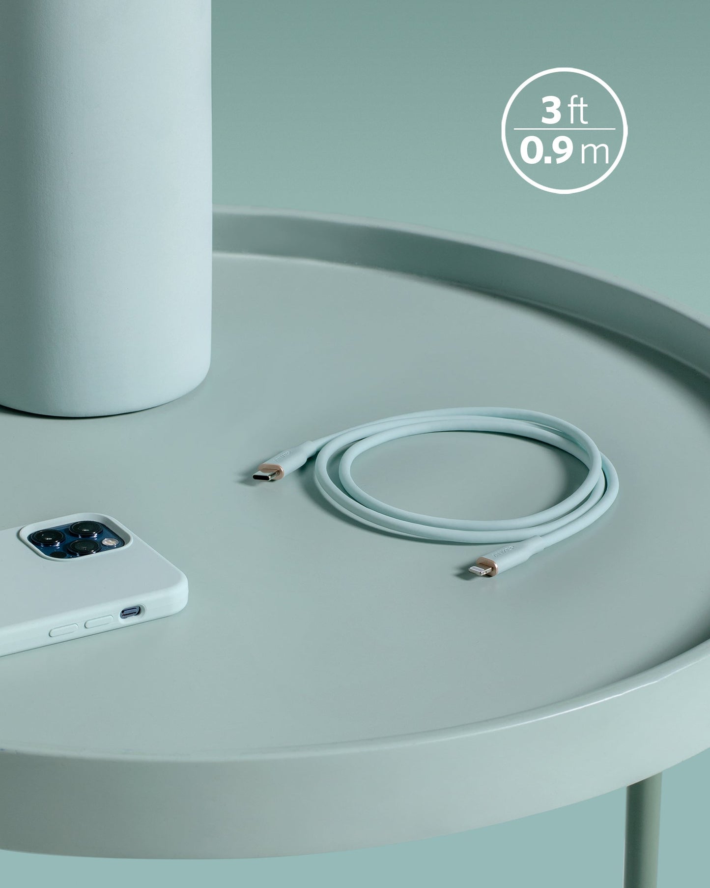 A coiled white Anker 641 USB-C to Lightning Cable (Flow, 3 ft Silicone) rests on a round light gray table beside a white triple-lens smartphone, with a large cylinder and “3 ft / 0.9 m” text adding to the sleek, minimal scene.