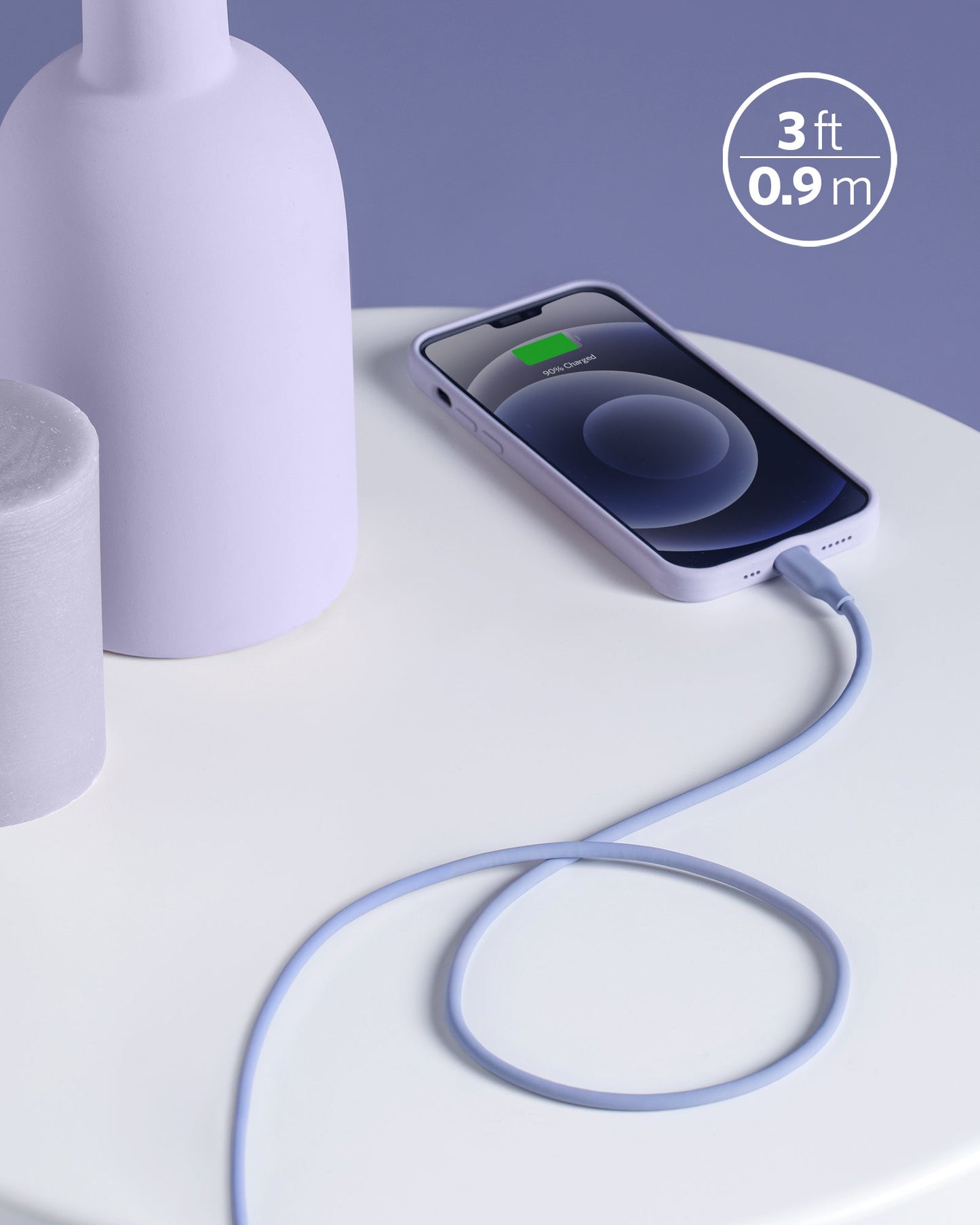 A smartphone shows a charging icon while connected by an Anker 641 USB-C to Lightning Cable (Flow, 3 ft Silicone) in light purple on a white round table. Nearby are a matte lavender bottle and cylinder, all in soft pastel tones. Brand: Anker.
