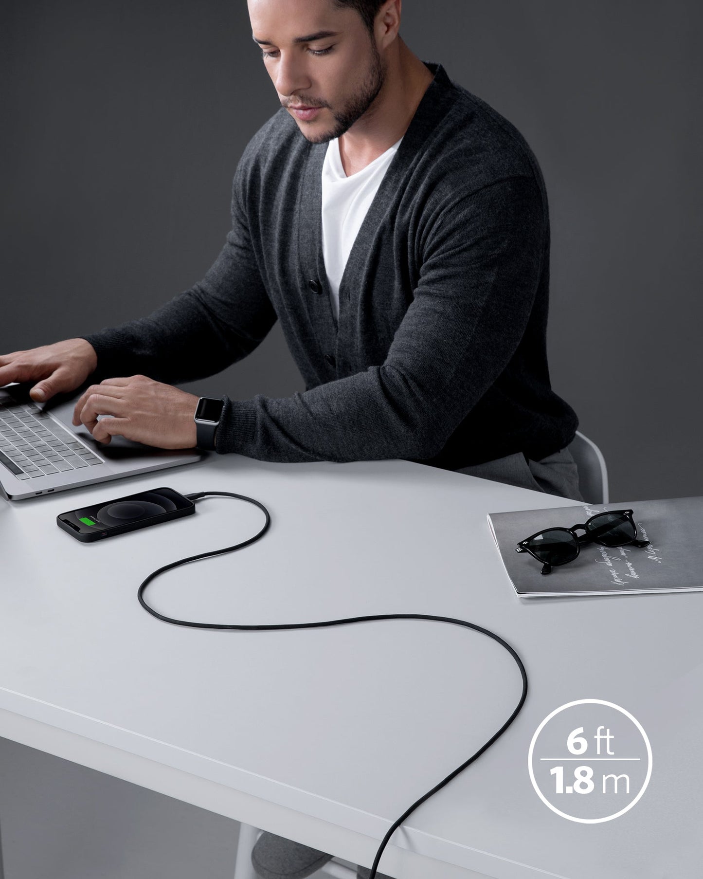 A man uses a laptop at a white desk with an Anker 641 USB-C to Lightning Cable (Flow, 3 ft Silicone) plugged into a device. The silicone cable trails toward the camera. Nearby are sunglasses, a gray notebook, and “3 ft” text in the corner.