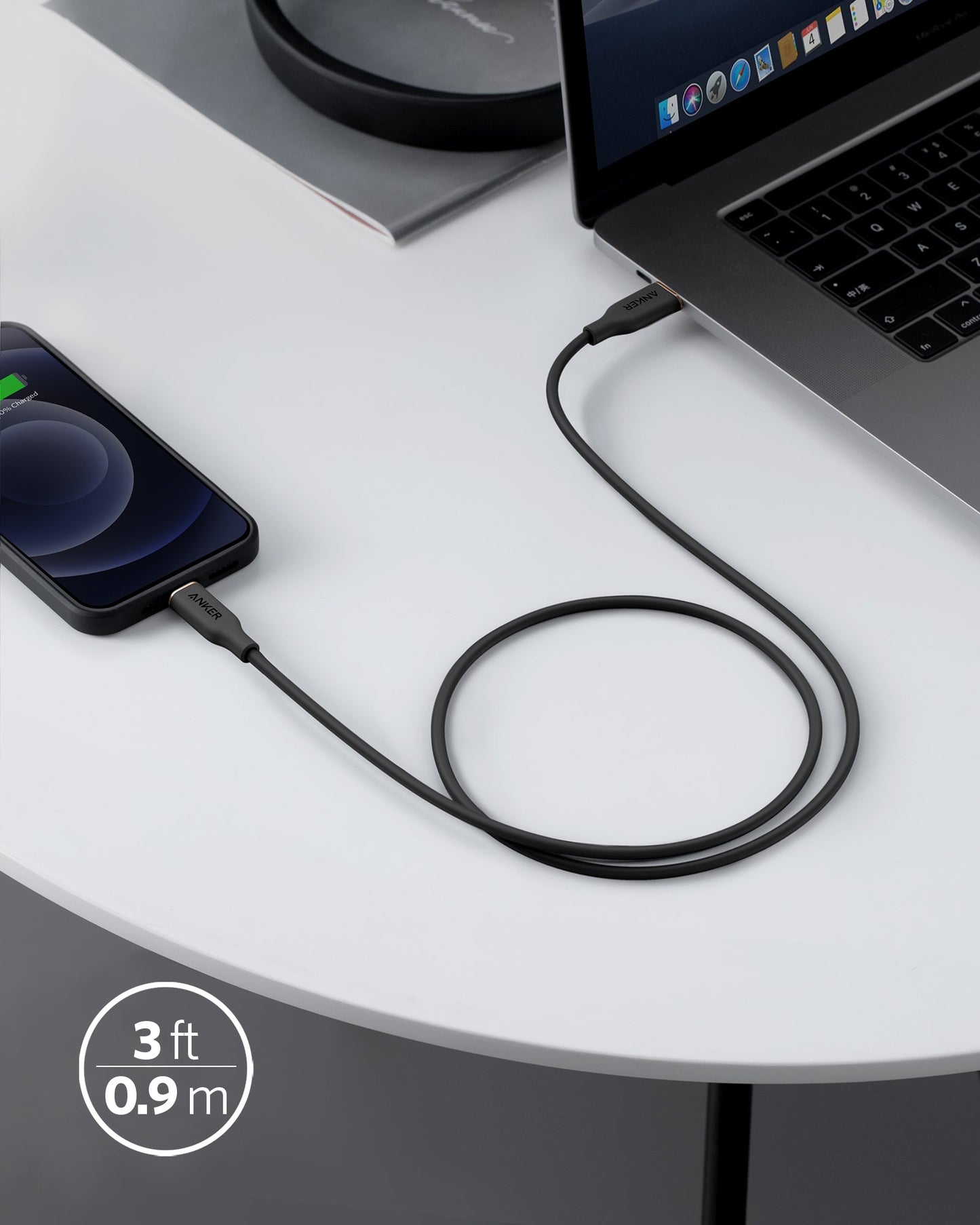 An Anker 641 USB-C to Lightning Cable (Flow, 3 ft Silicone) in black connects an iPhone with a charging symbol to a MacBook on a white round table. A “3 ft / 0.9 m” label and a closed notebook are also partially visible.