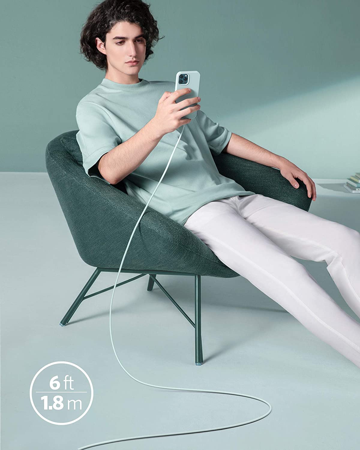 A person with wavy hair sits in a dark green modern chair, holding their smartphone connected to an Anker 641 USB-C to Lightning Cable (Flow, 3 ft Silicone) by Anker. The silicone cable stretches across a light floor and pale green background.