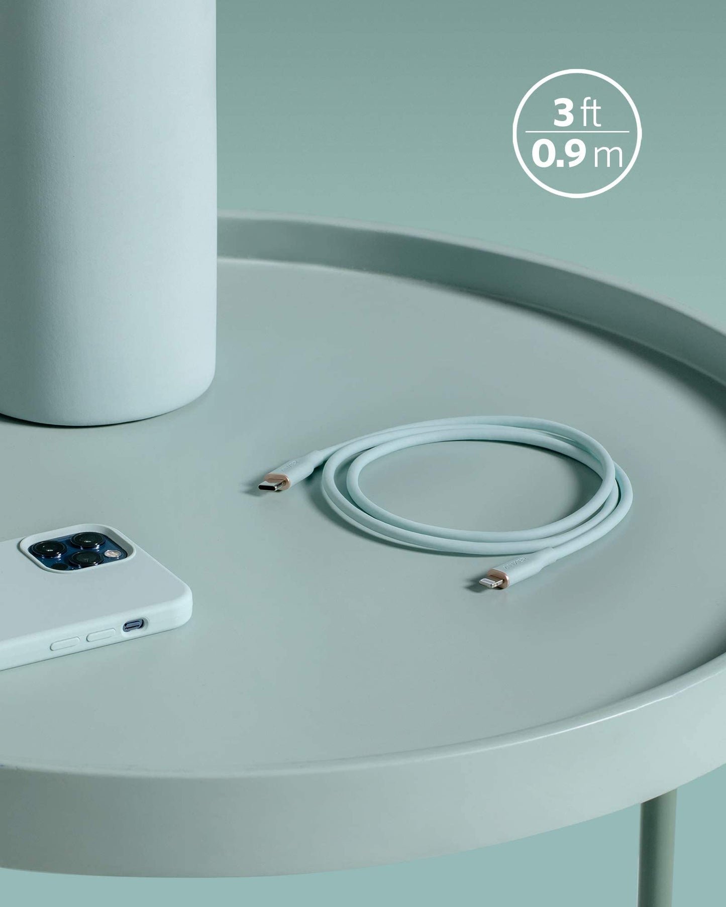 A coiled Anker 641 USB-C to Lightning Cable (Flow, Silicone) in light gray rests on a round, pale green table next to a smartphone and cylindrical item. “3 ft 0.9 m” appears in the corner. The scene is minimalist with soft colors.
