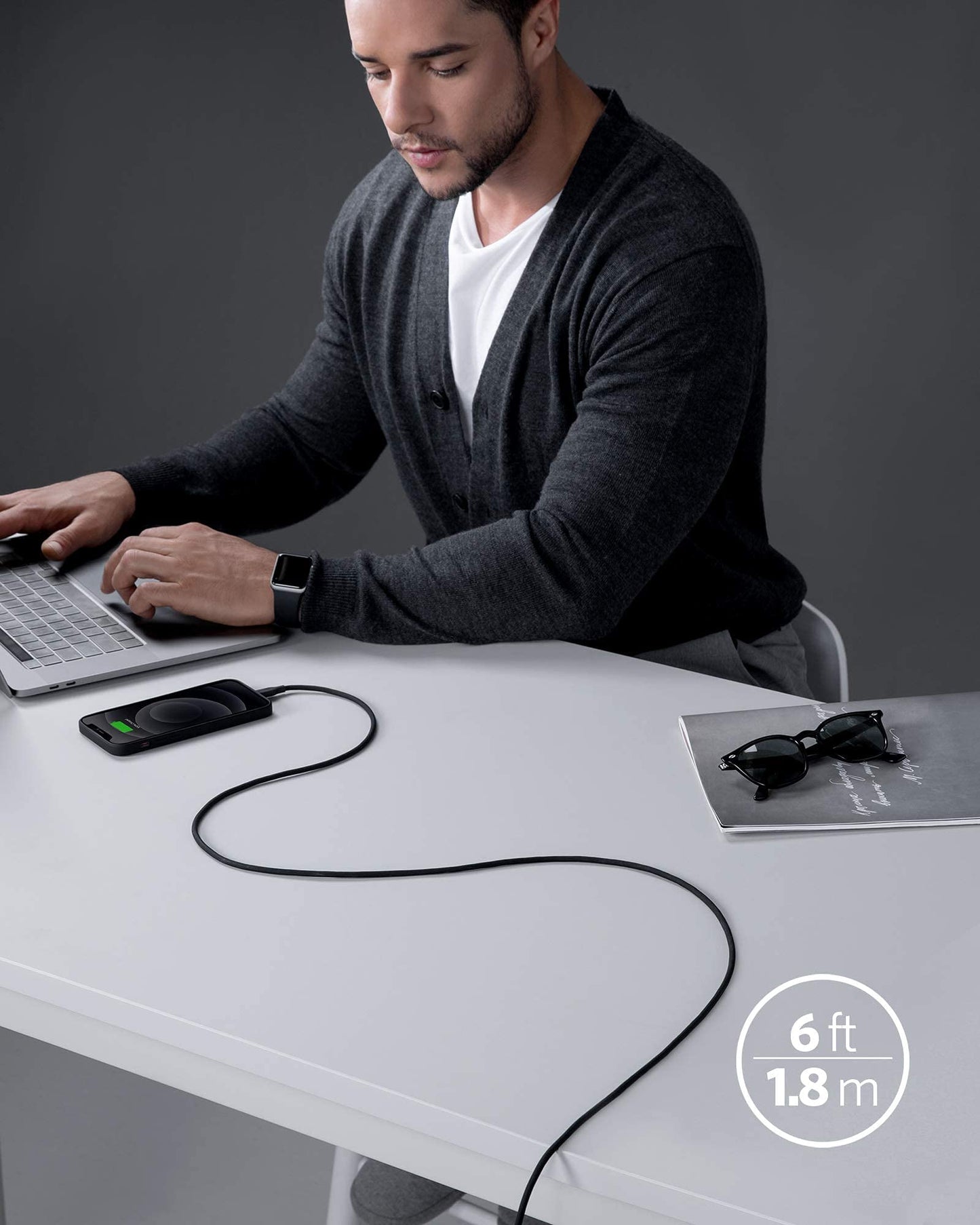 A man in a dark cardigan works on a laptop at a white desk. An Anker 641 USB-C to Lightning Cable (Flow, Silicone) connects his smartphone. Sunglasses and a closed notebook lie nearby against the dark, minimalist background.