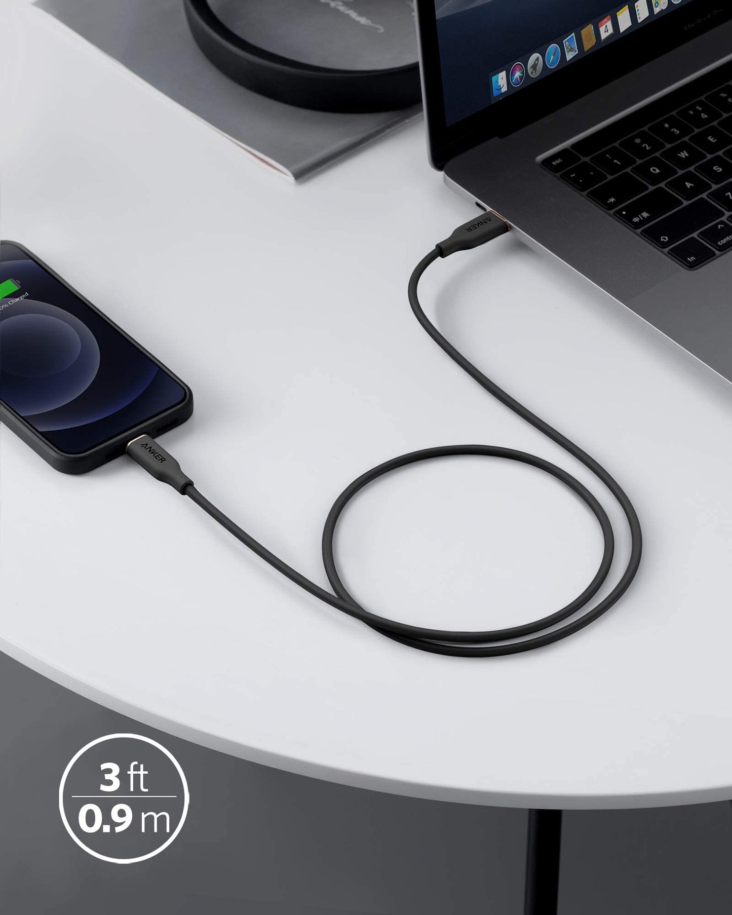An Anker 641 USB-C to Lightning Cable (Flow, Silicone) in black connects a smartphone showing a charging symbol to a laptop on a white round table labeled 3 ft / 0.9 m, with part of a magazine and lamp visible in the background.
