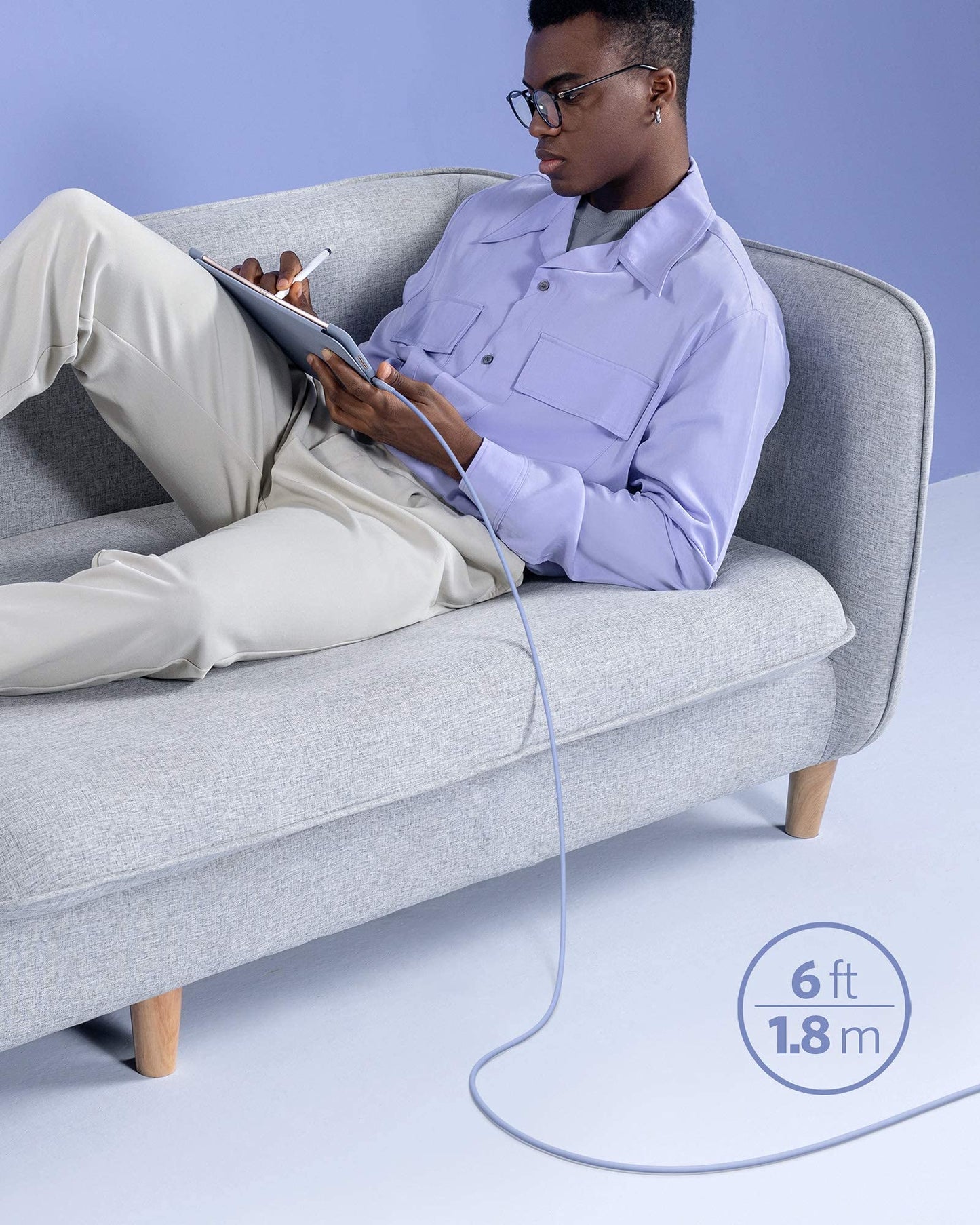 A person in light pants and a lavender shirt lounges on a light gray sofa, charging their tablet with the Anker 641 USB-C to Lightning Cable (Flow, Silicone), a 6 ft MFi certified cable in pale purple. "Anker" branding appears on the background.