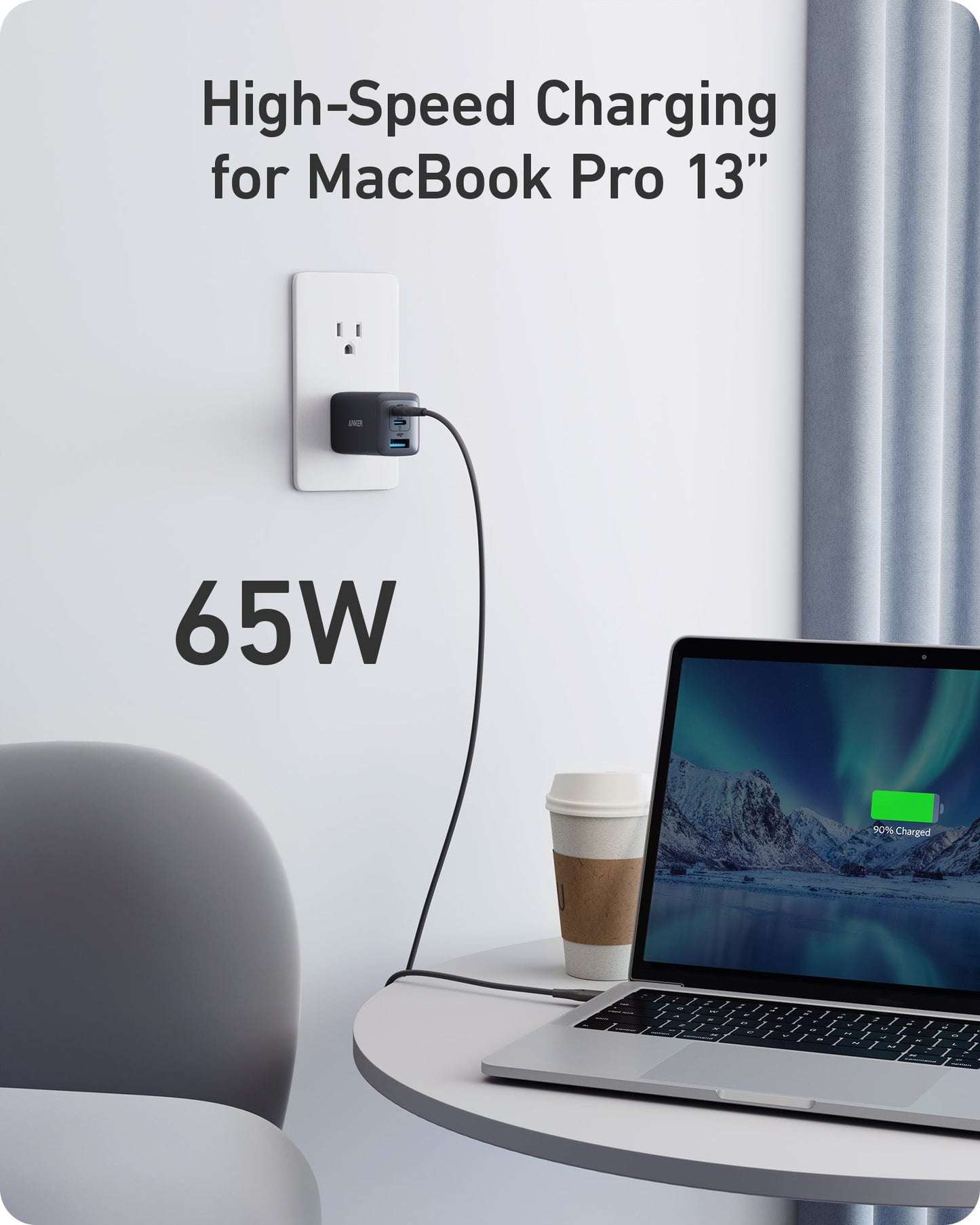 An Anker 735 Charger (Nano II 65W) is plugged into a wall outlet, fast charging a MacBook Pro 13" to 100% on a white round table. A coffee cup, gray chair, and curtain can also be seen.