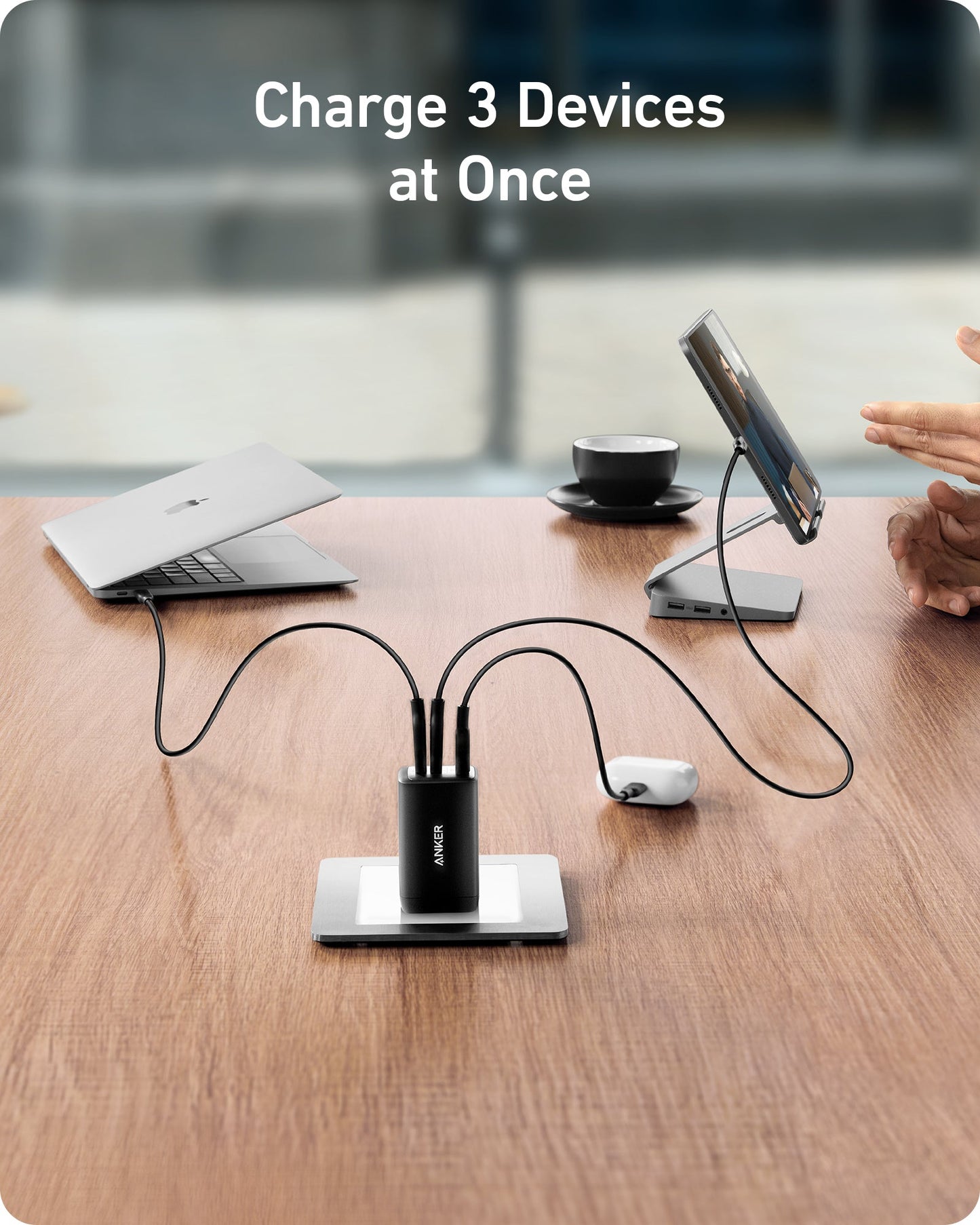 A compact black Anker 735 Charger (Nano II 65W) with GaN II fast charges a laptop, tablet, and mouse at once on a wooden table. Two people interact nearby, with a blurred window and coffee cup in the background.