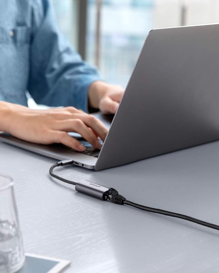 A person in a blue shirt types on a silver laptop at a gray table. An Anker <b>PowerExpand</b> USB-C to Gigabit Ethernet Adapter with a black cable is connected, while a blurry glass of water sits in the left foreground.