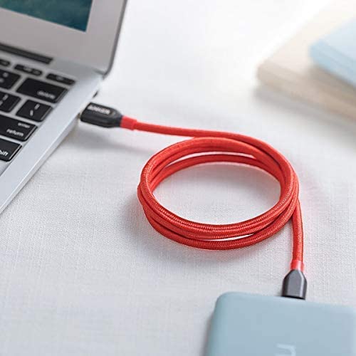 A red Anker Powerline+ USB C to USB 3.0 Cable connects a silver laptop to a small gray device on a white surface, loosely coiled, with part of a light blue notebook visible in the upper right background.